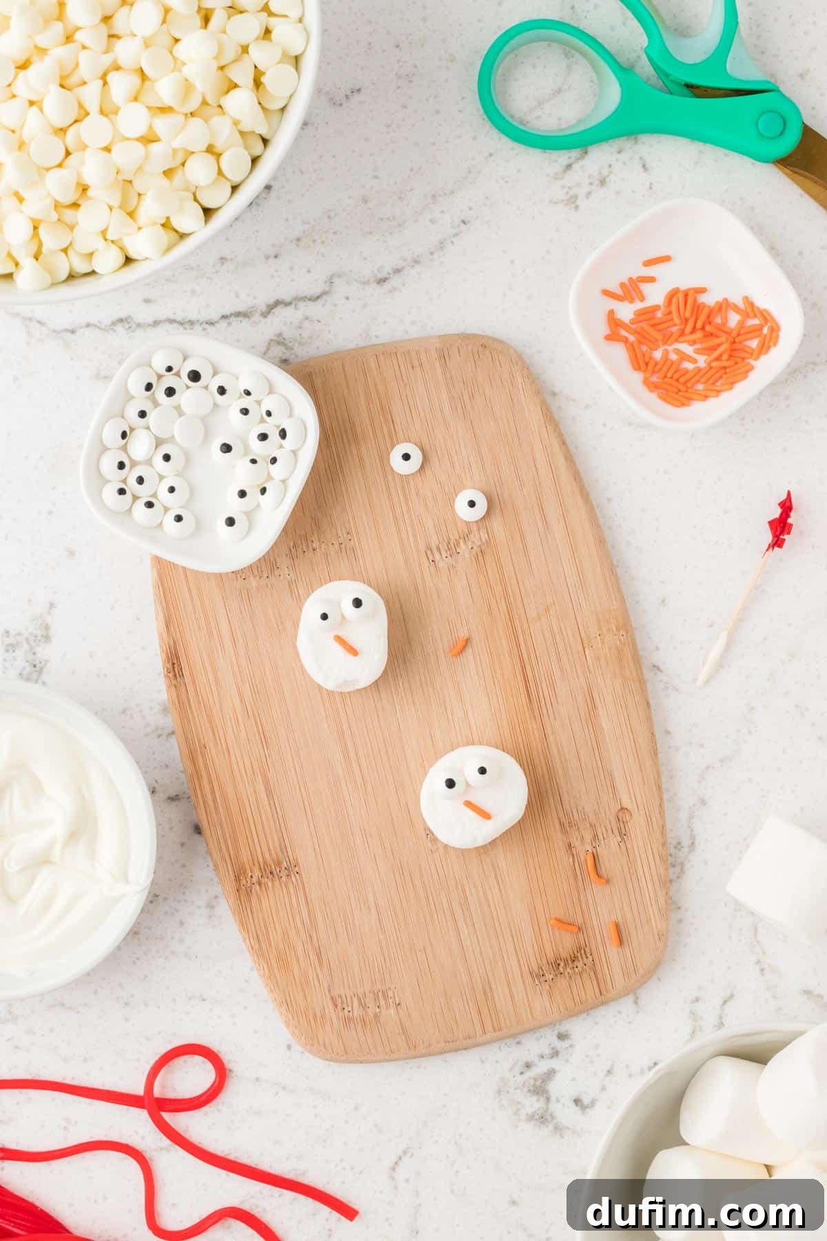 A step-by-step visual guide showing how to attach candy eyeballs and an orange jimmy nose to marshmallow halves using melted chocolate, creating adorable snowman heads.