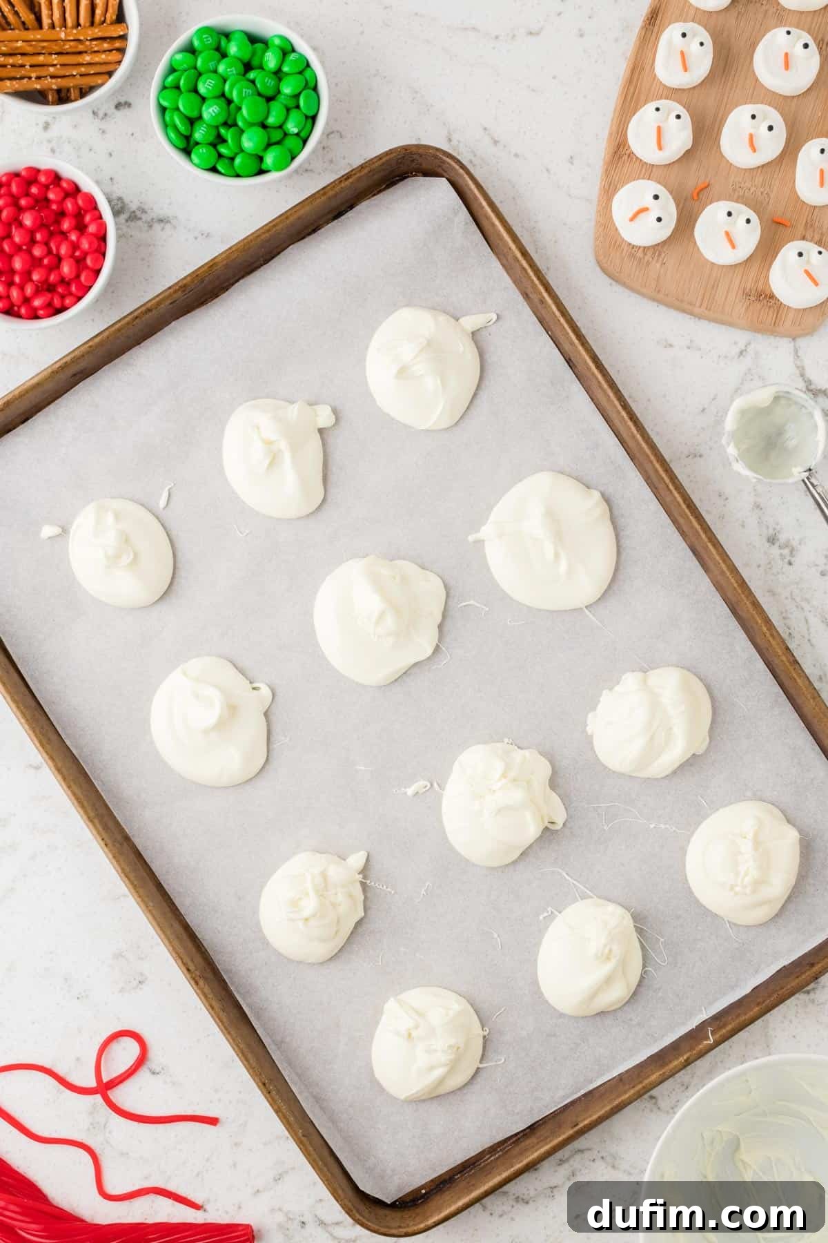 Evenly spaced dollops of melted white chocolate arranged in a grid pattern on parchment paper, ready for snowman assembly.