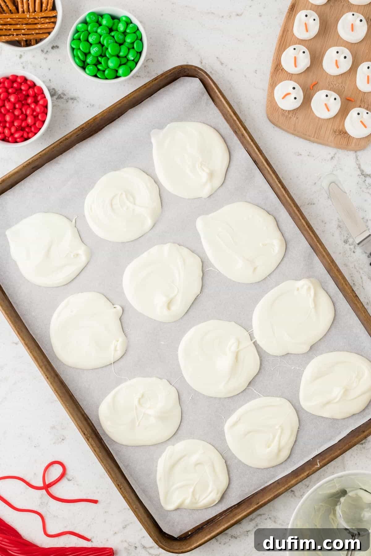 Melted white chocolate dollops carefully spread into circular shapes on parchment paper, forming the base for the snowman bark.