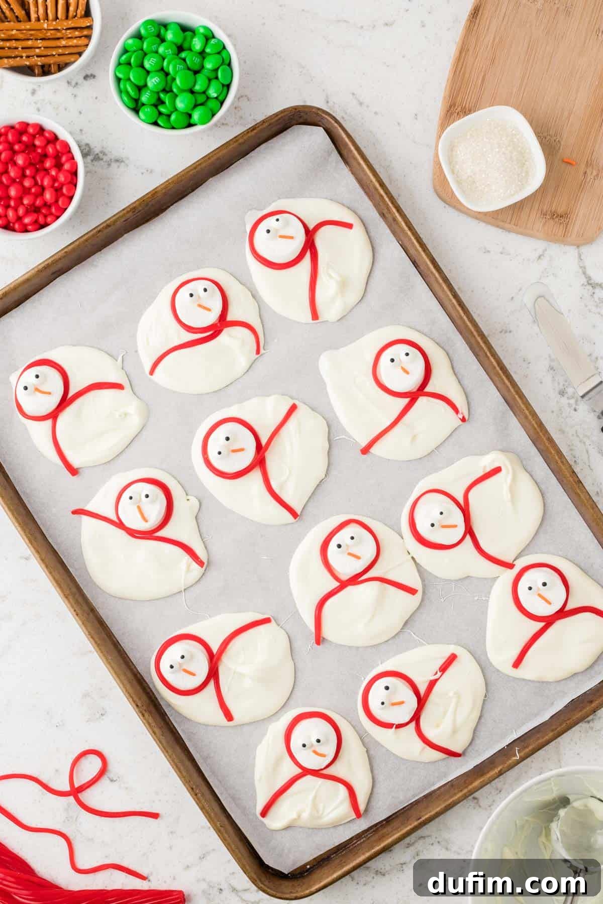 Partially assembled snowman bark with marshmallow heads on white chocolate dollops, as Twizzlers are being wrapped to form scarves.