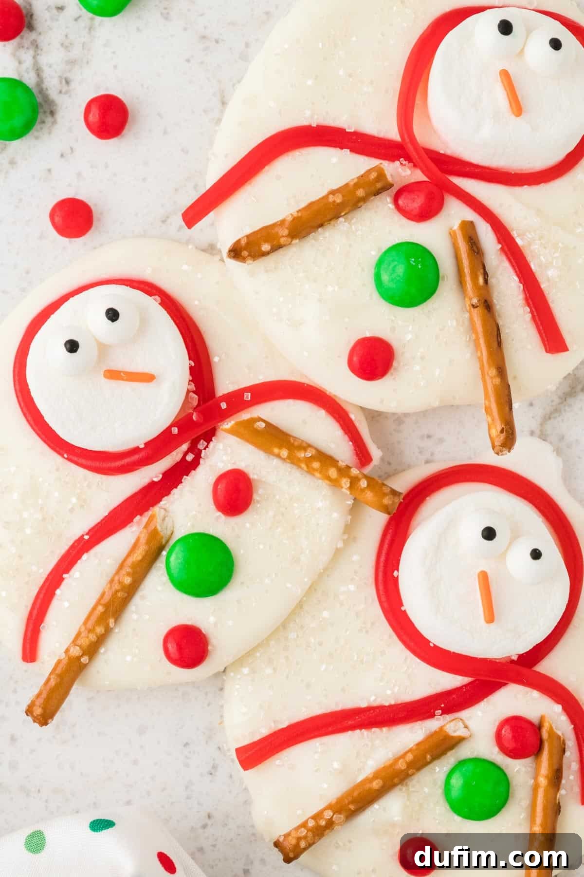 A close-up view of melted snowman bark, highlighting the intricate details of the marshmallow snowmen with their candy eyes, orange noses, colorful Twizzler scarves, pretzel arms, and candy buttons, all nestled on a bed of white chocolate bark.
