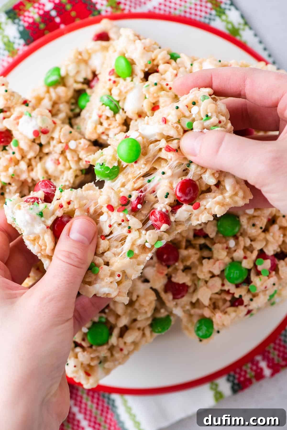 A hand reaching for two colorful Christmas rice krispie treats from a white plate, ready to be enjoyed.