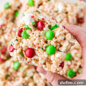 A hand gently holding a soft, chewy Rice Krispie treat adorned with festive red and green M&Ms and sprinkles.