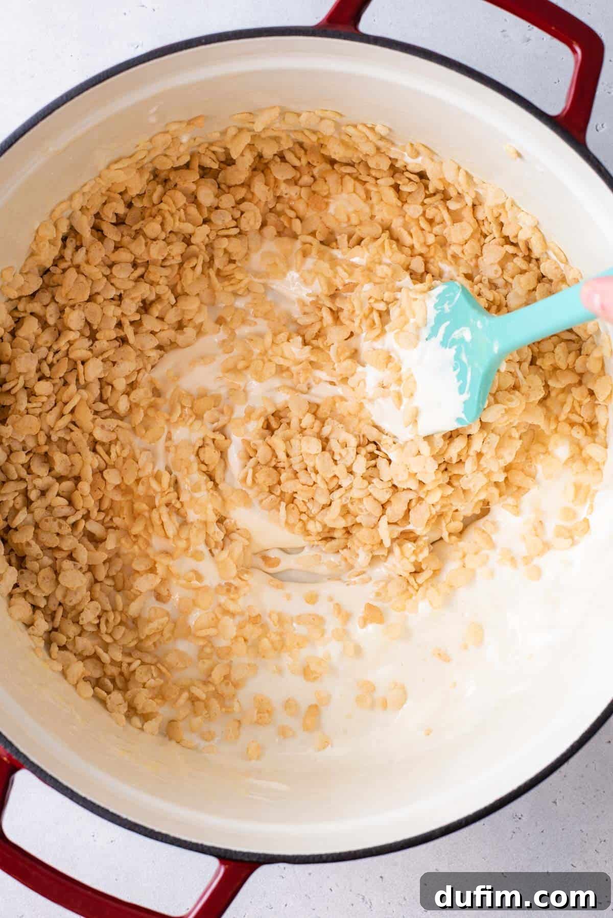 A blue spatula expertly stirring Rice Krispie cereal into the golden, melted marshmallow mixture in a large saucepan.