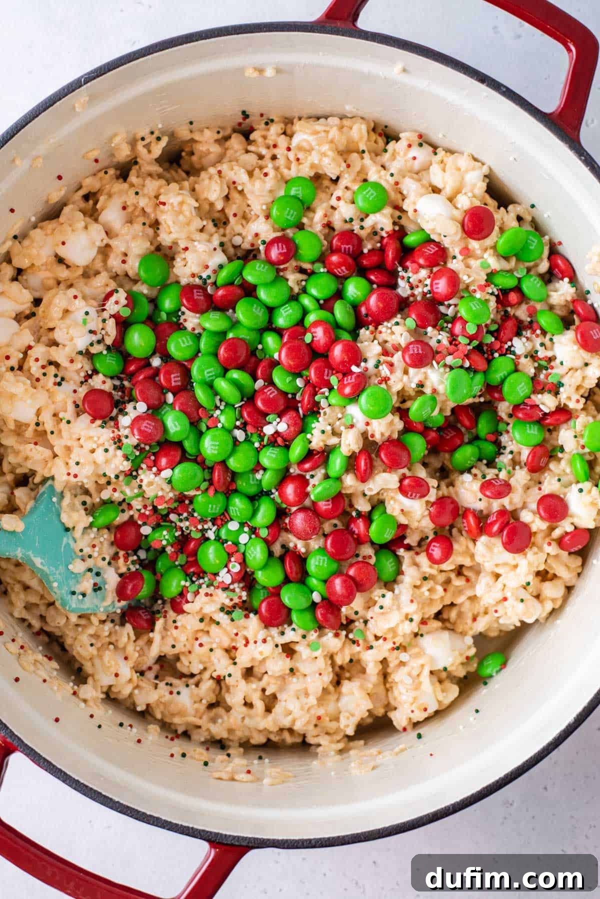 Vibrant red and green Christmas M&Ms being gently stirred into the sticky Rice Krispie mixture.