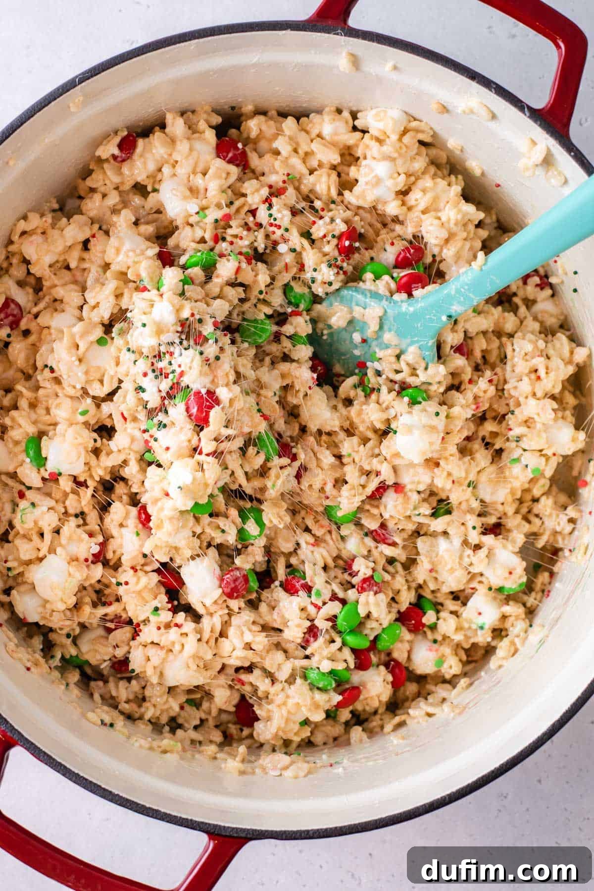 A spatula stirring Christmas M&Ms and colorful sprinkles into the Rice Krispie treat mixture for a festive finish.