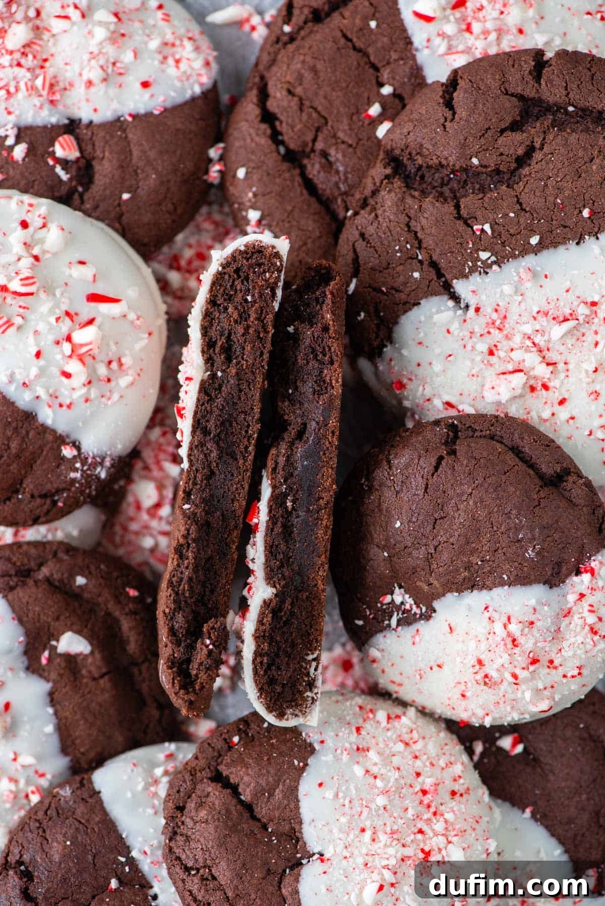 These Chocolate Candy Cane Cookies feature soft, chewy chocolate cookies dipped in white chocolate and sprinkled with crushed candy canes. A chocolate candy cane cookie neatly cut in half, revealing its soft, fudgy interior, placed on top of a stack of other festive cookies.