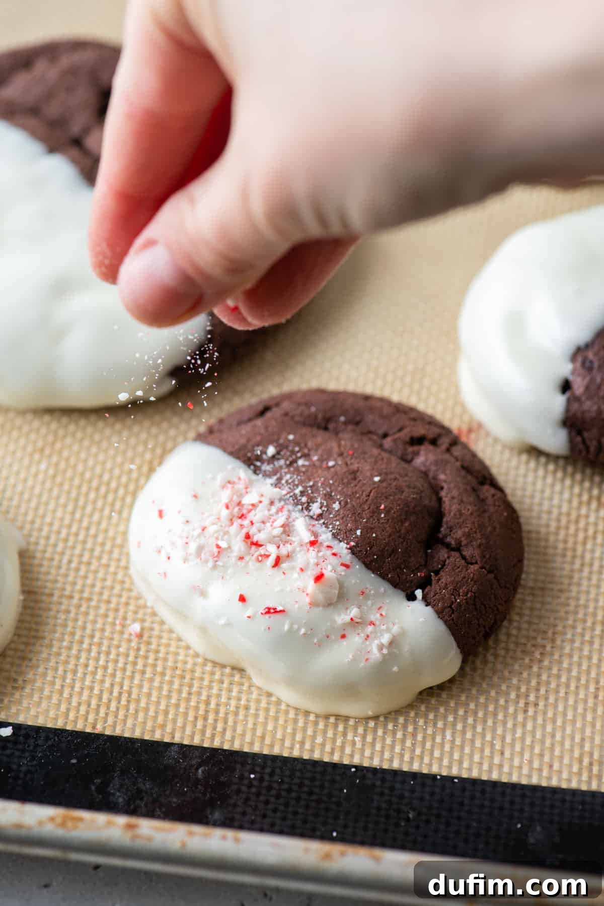 These Chocolate Candy Cane Cookies feature soft, chewy chocolate cookies dipped in white chocolate and sprinkled with crushed candy canes. A hand gently sprinkling shimmering crushed candy canes over a freshly white chocolate-dipped cookie, creating a festive finish.