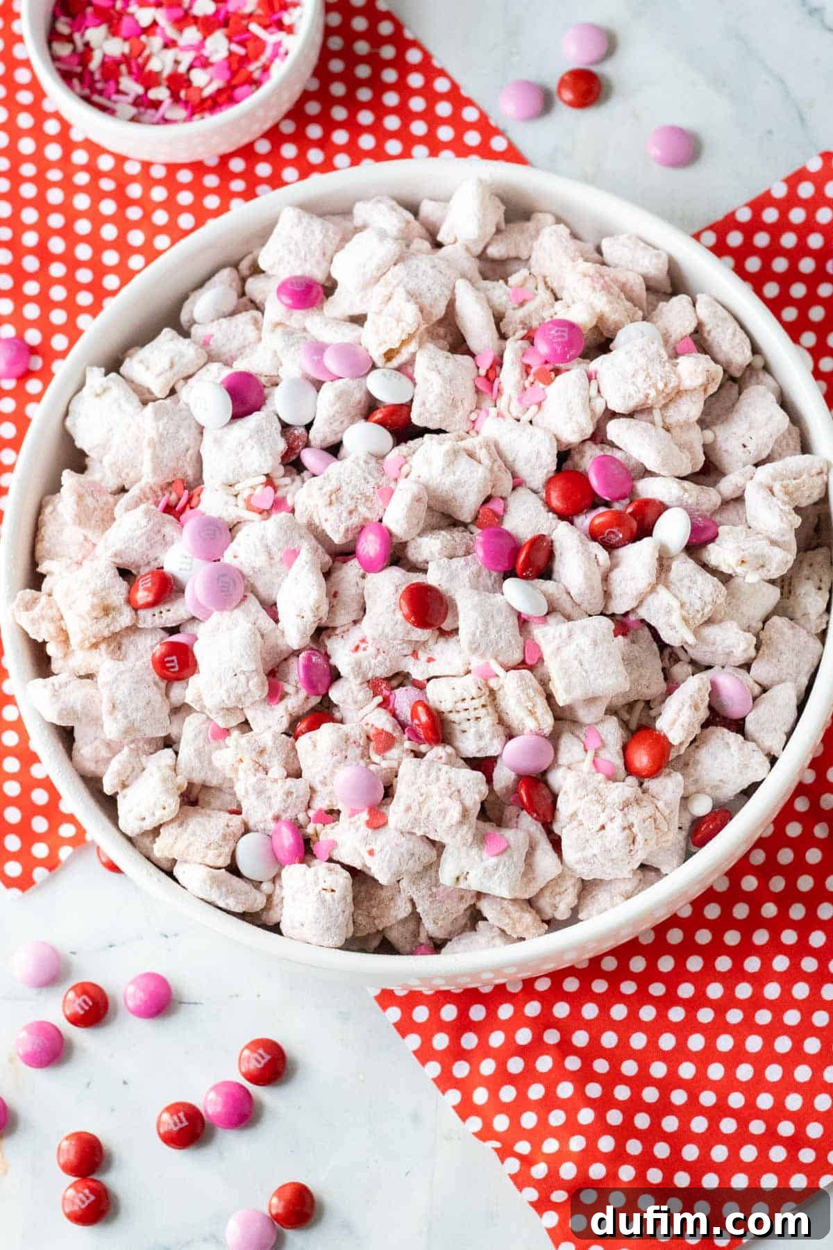 Valentine's Day Puppy Chow in a white bowl sitting on top of a white counter and red napkins with white polka dots on them.