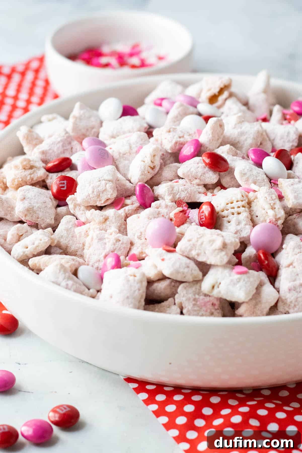 Valentine's Day Puppy Chow treat in a white bowl sitting on top of red and white napkin with pink & red m&ms sprinkled around it.