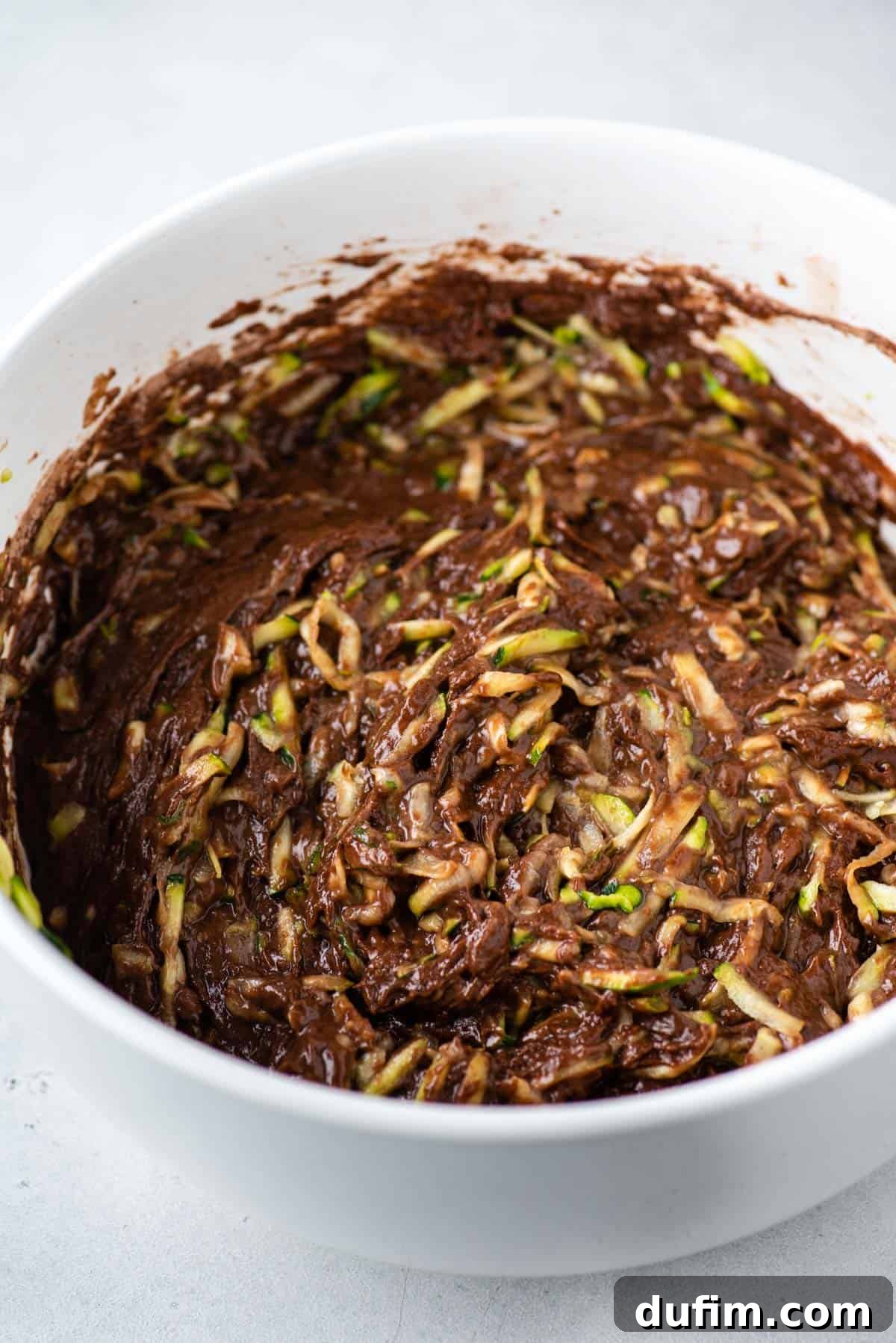 These Chocolate Zucchini Muffins are rich, moist, and oh-so-good. The zucchini adds moisture while cocoa powder adds chocolate decadence. Shredded zucchini being gently mixed into a rich chocolate muffin batter in a large bowl.