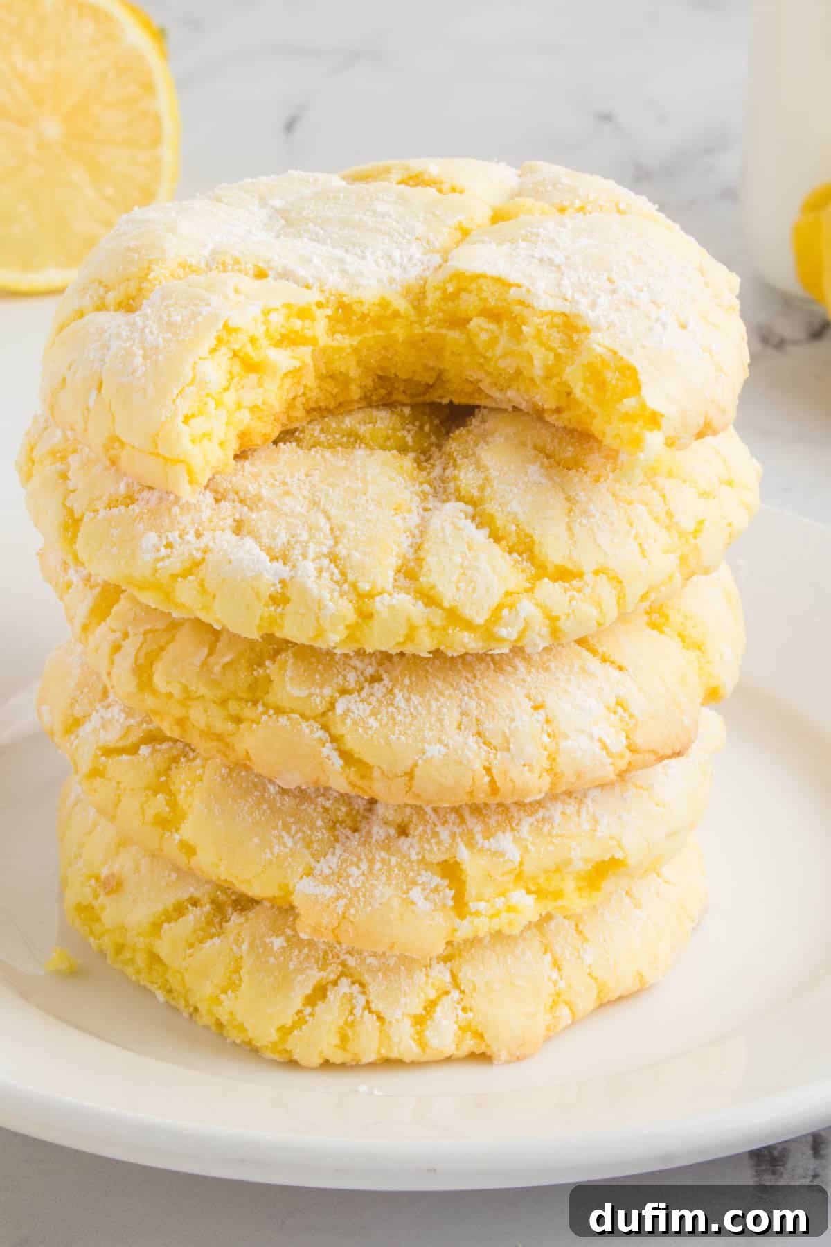 Stack of four moist lemon cake mix cookies on a white plate, garnished with a fresh lemon slice and mint sprig, showing their crinkled powdered sugar coating.