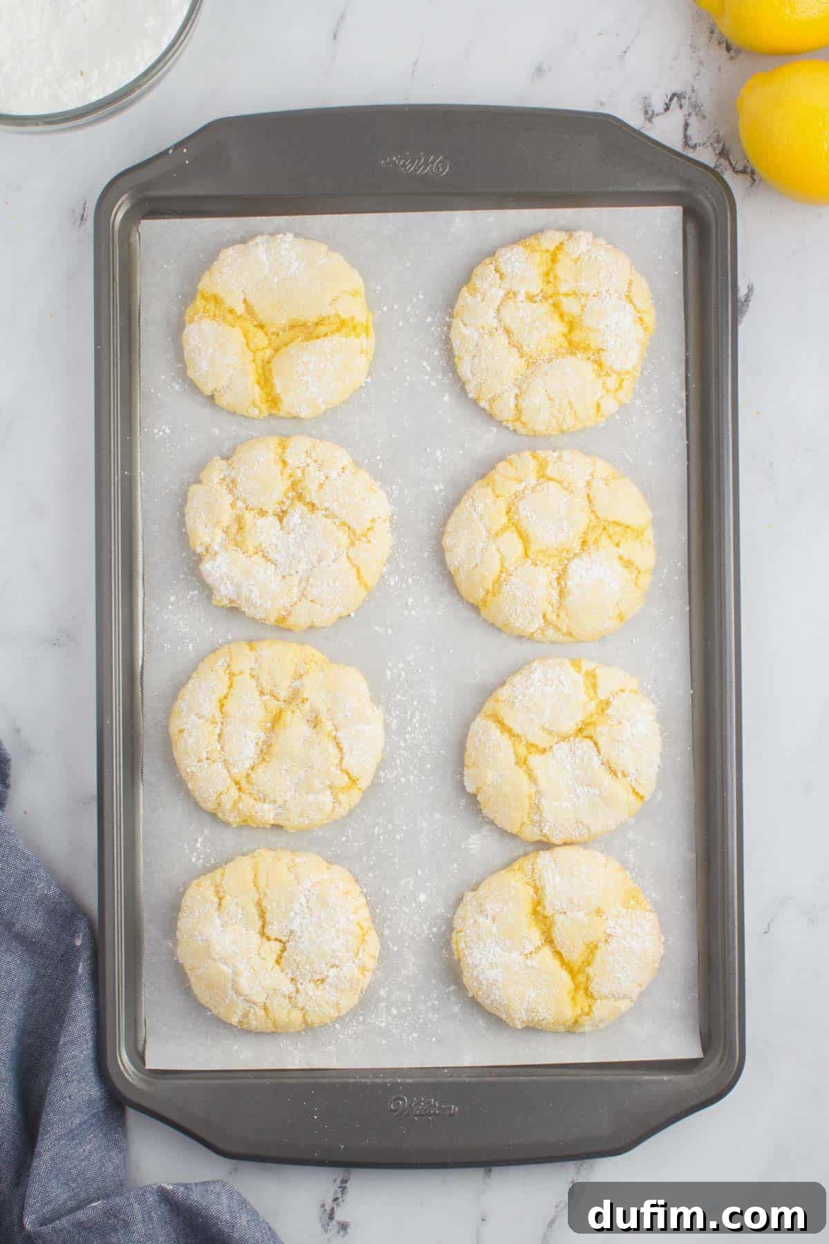Eight golden lemon cake mix cookies cooling on a parchment-lined baking sheet on a granite countertop, ready to be enjoyed.