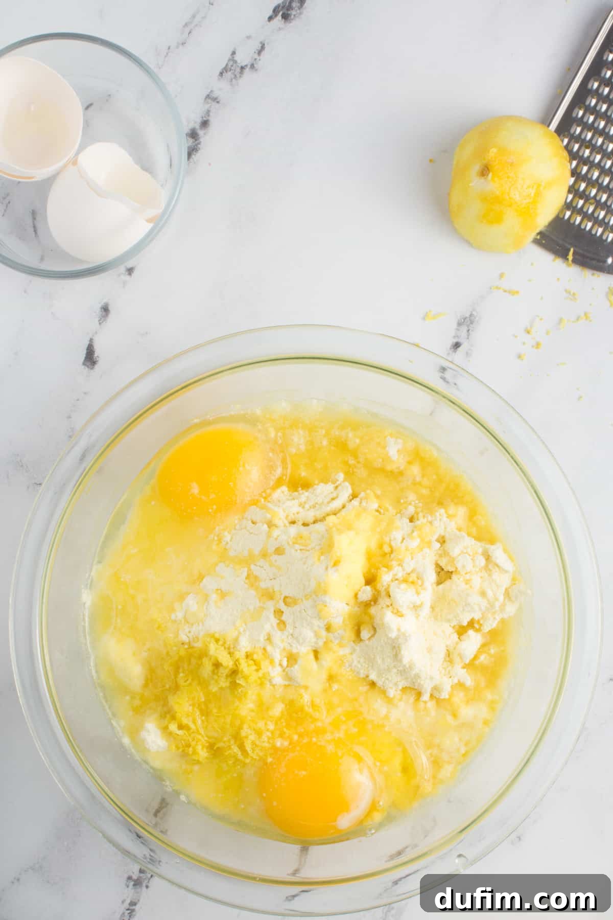 Ingredients for lemon cake mix cookies partially mixed in a large glass bowl on a kitchen counter, surrounded by fresh lemons, a grater, and empty eggshells.