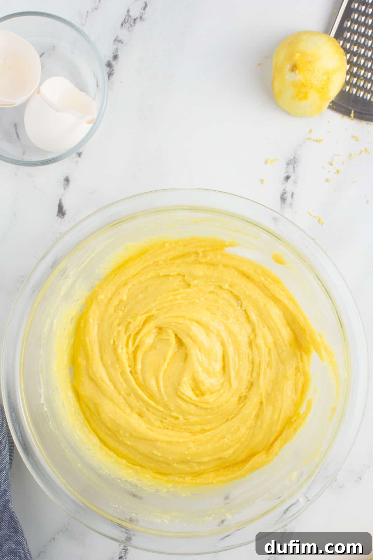 Smooth, sticky lemon cookie dough in a clear glass mixing bowl resting on a granite countertop, ready for chilling.