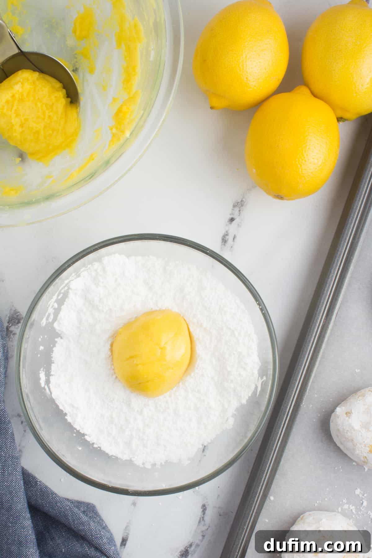 A baking sheet lined with parchment paper, featuring a bowl of powdered sugar, a bowl of lemon cookie dough, and fresh lemons and a grater on the side.