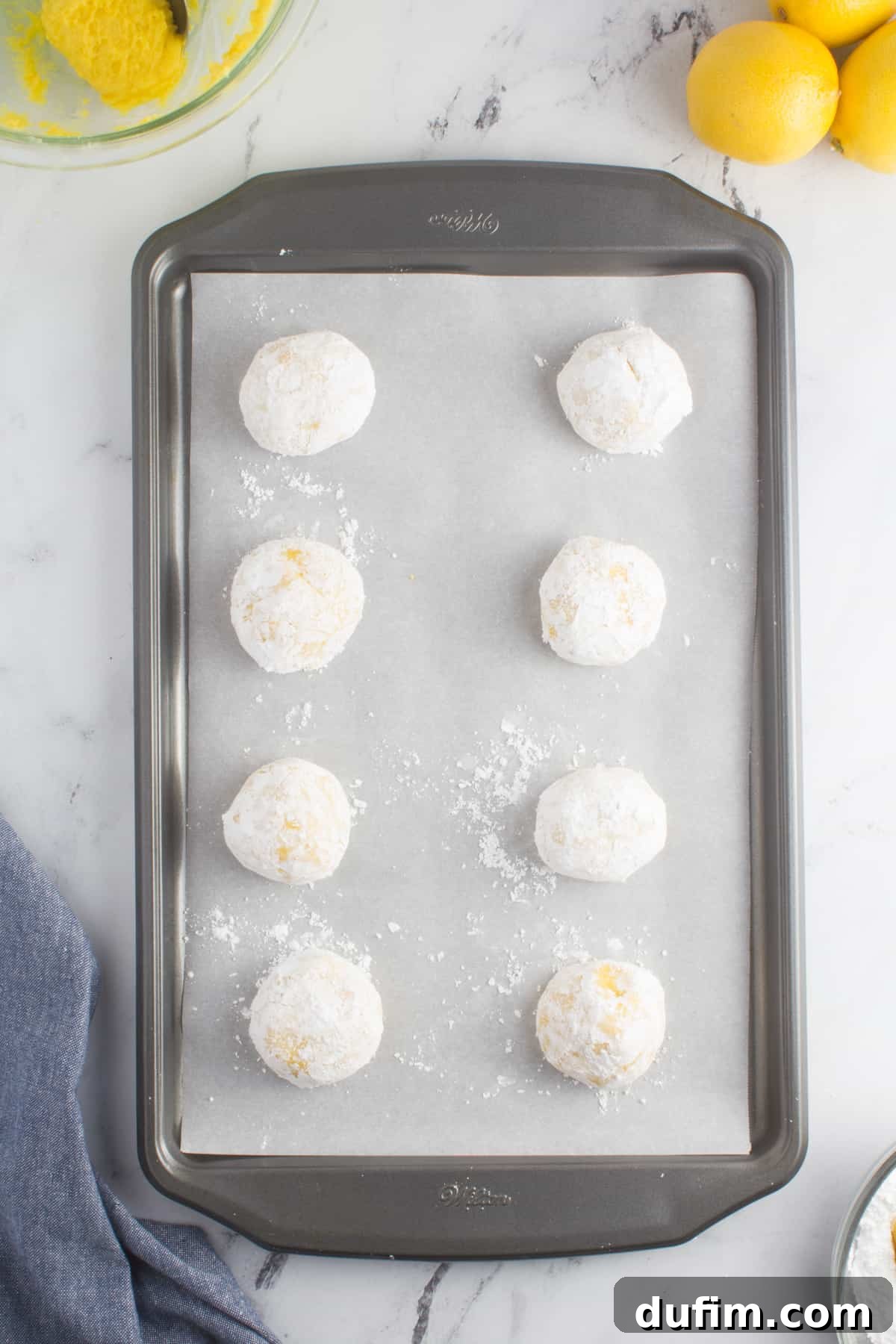 Lemon cookie dough balls generously coated in powdered sugar, arranged on a baking sheet lined with parchment paper, ready for the oven.