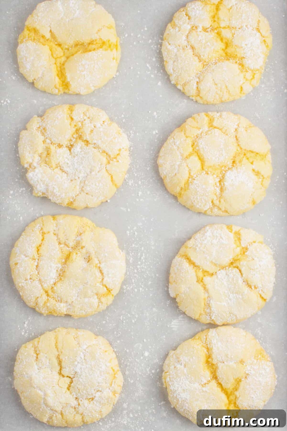Eight perfectly baked lemon cake mix cookies, golden and puffy with their signature crinkled powdered sugar tops, cooling on a parchment-lined baking sheet.