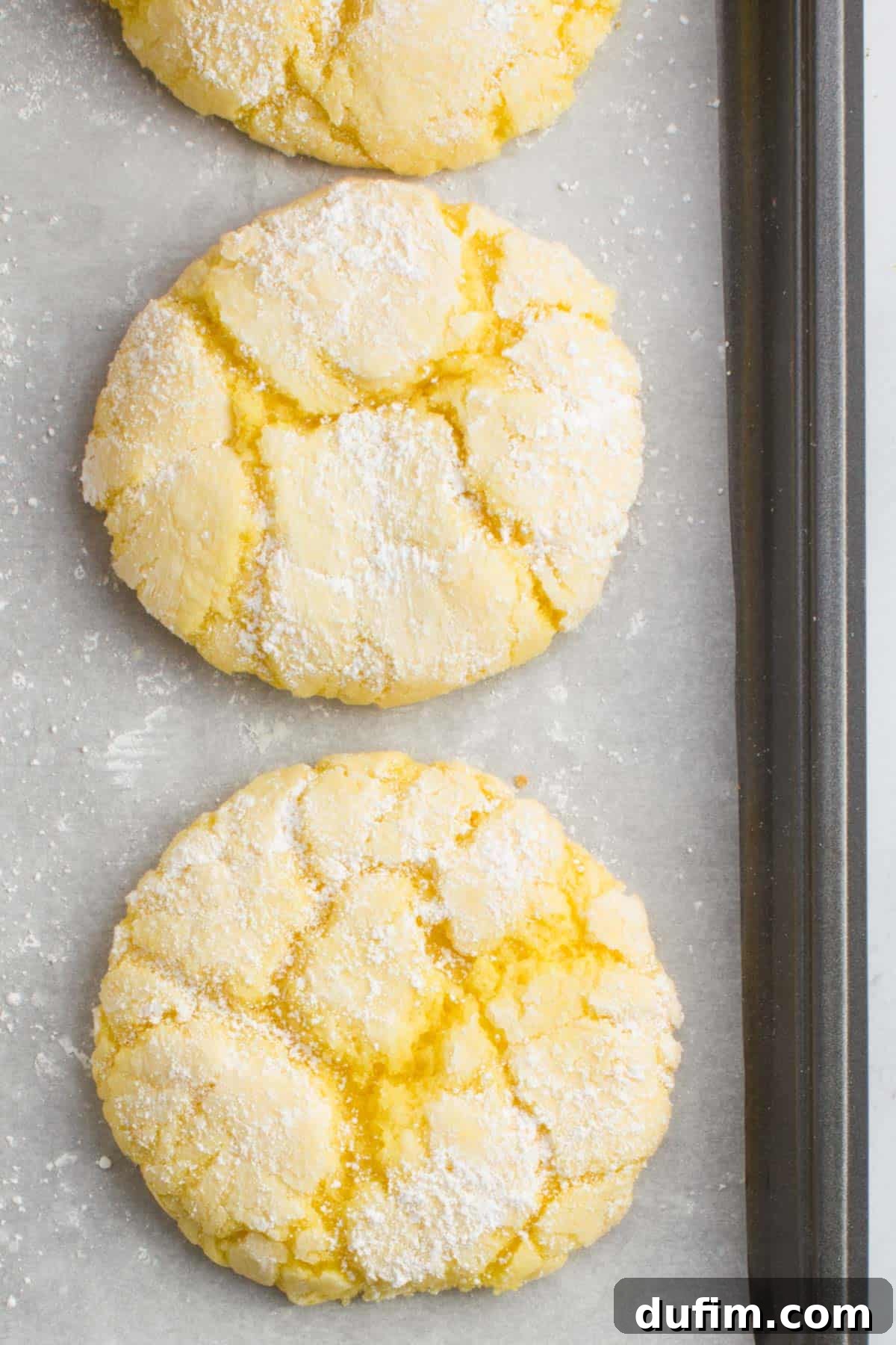 Two freshly baked lemon cake mix cookies cooling on a parchment-lined baking sheet, showcasing their fluffy texture and crinkled tops.