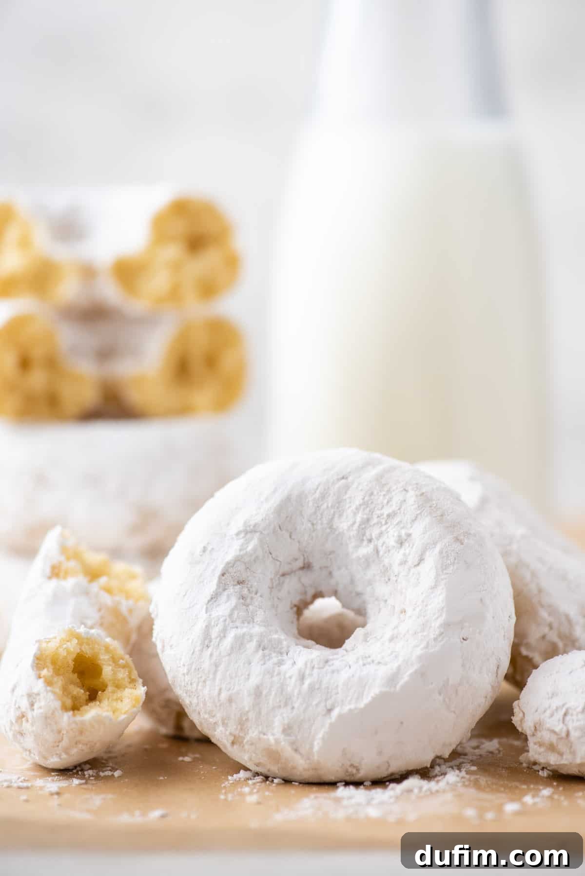Close-up of one whole and one half-eaten powdered sugar donut, showcasing their soft texture and generous sugar coating, resting on brown paper.