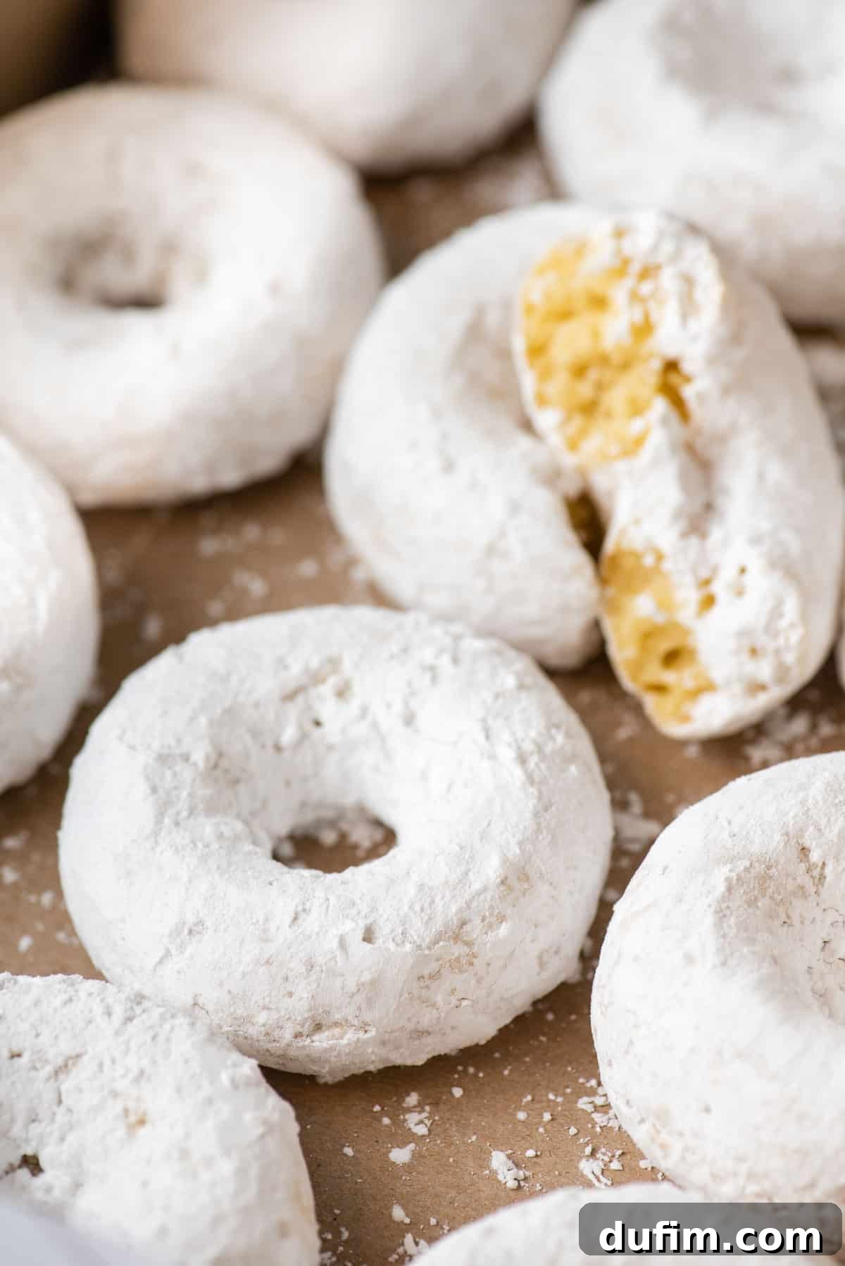 Close-up view of multiple powdered sugar donuts stacked together on brown paper, highlighting their delicate white coating and soft interior.