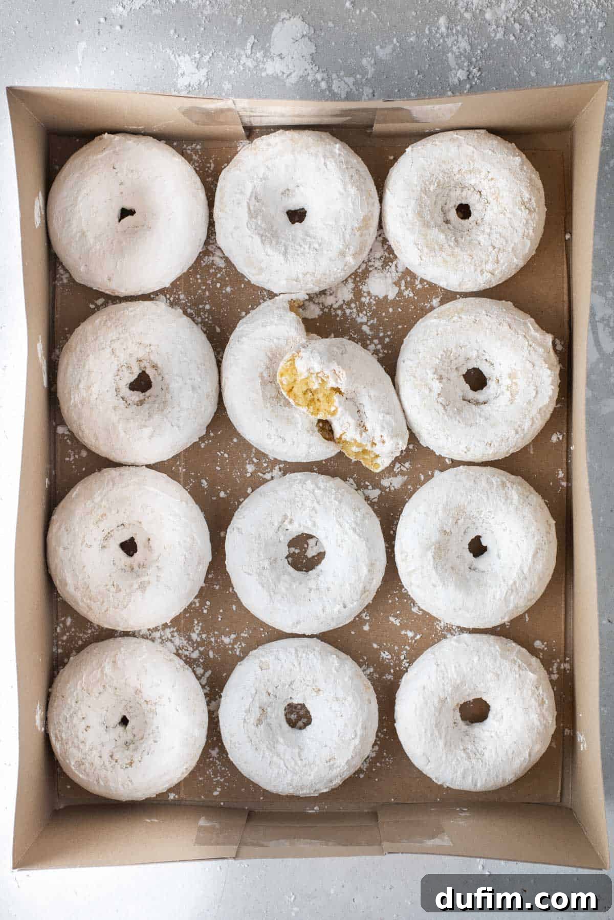Rows of freshly baked powdered sugar donuts neatly arranged in a bakery box on a countertop, ready to be served or shared.