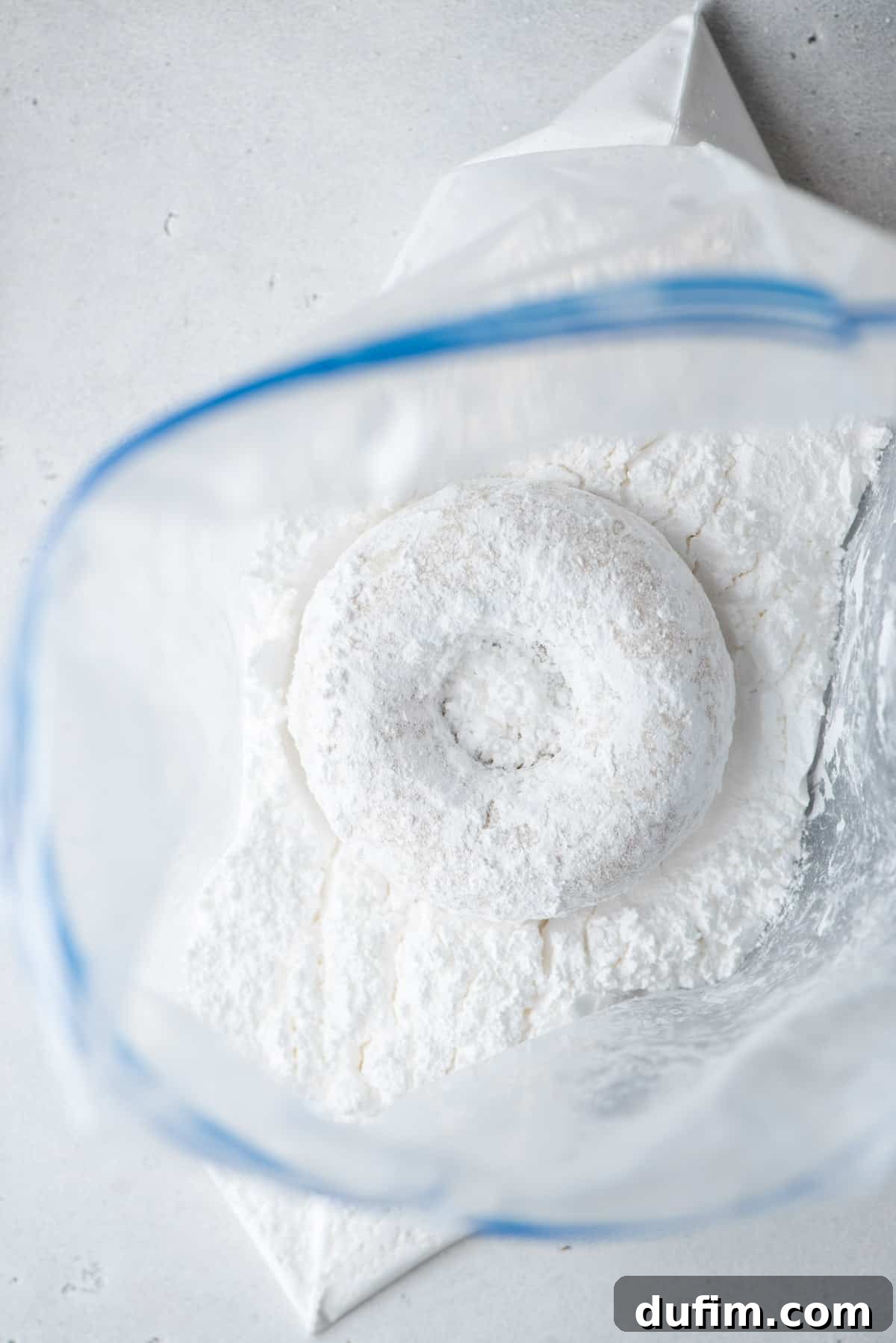 A baked donut being shaken in a plastic bag filled with powdered sugar, illustrating the coating process.