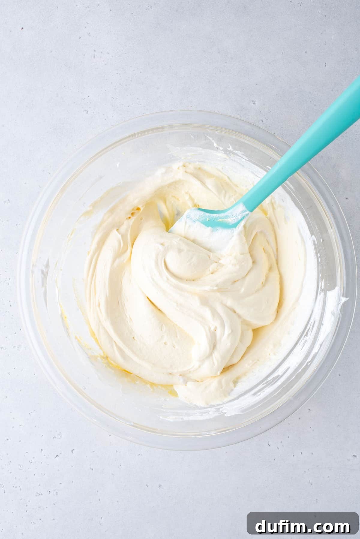 cool whip frosting in a glass bowl being stirred with a teal spatula