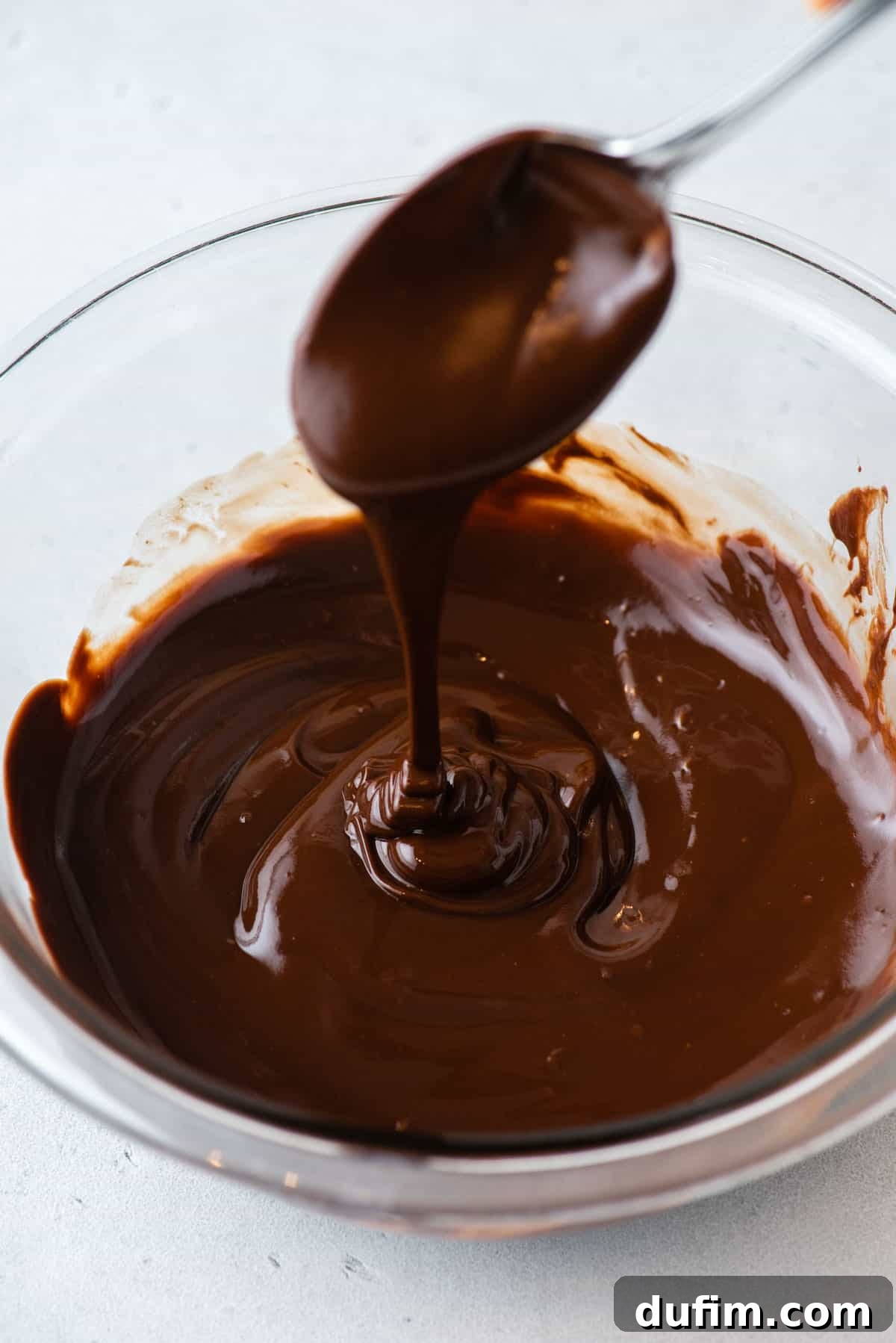 A clear glass bowl containing a pool of rich, melted chocolate glaze, ready for dipping donuts.