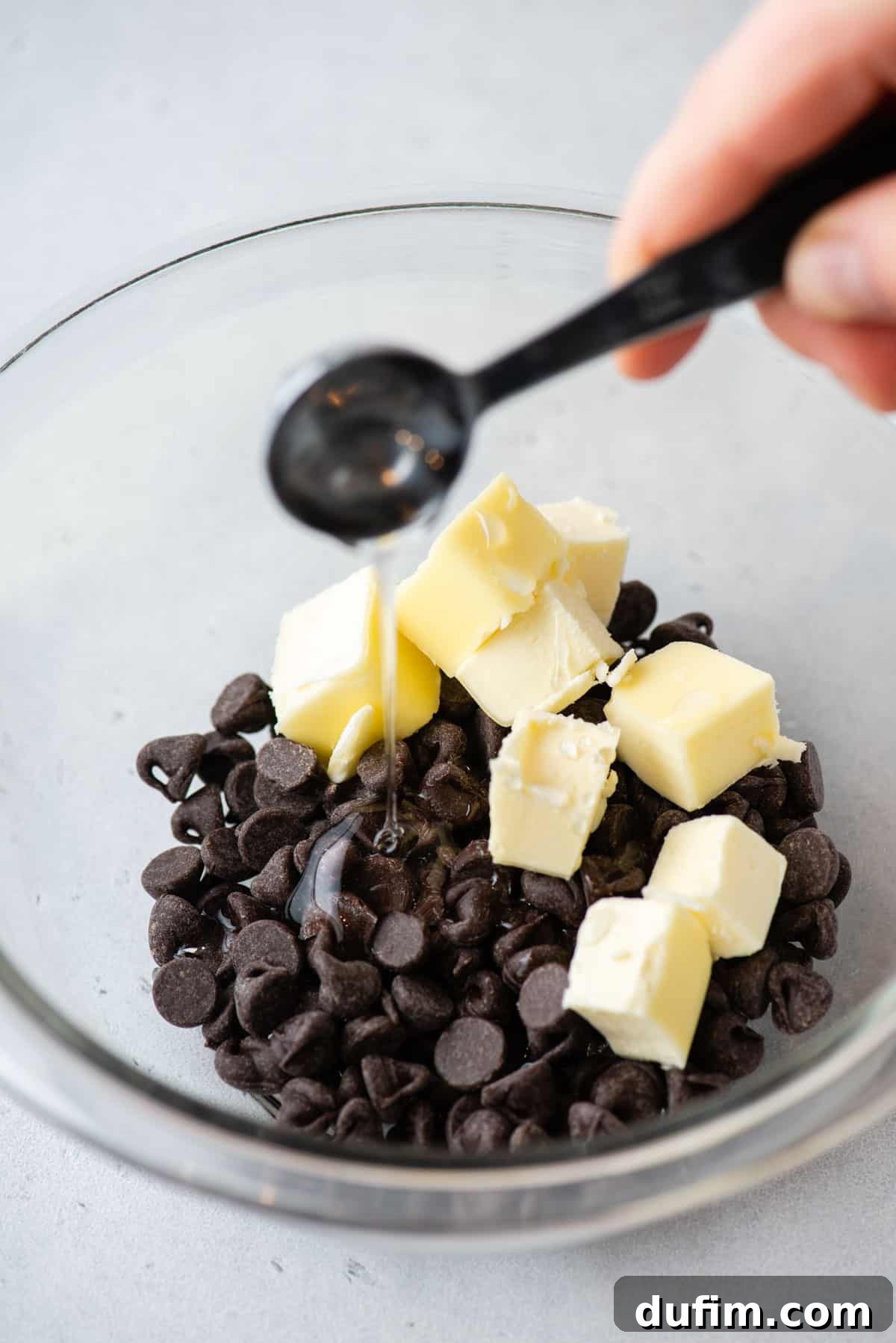 Semi-sweet chocolate chips and chunks of unsalted butter resting in a clear glass bowl, with light corn syrup being carefully poured in.