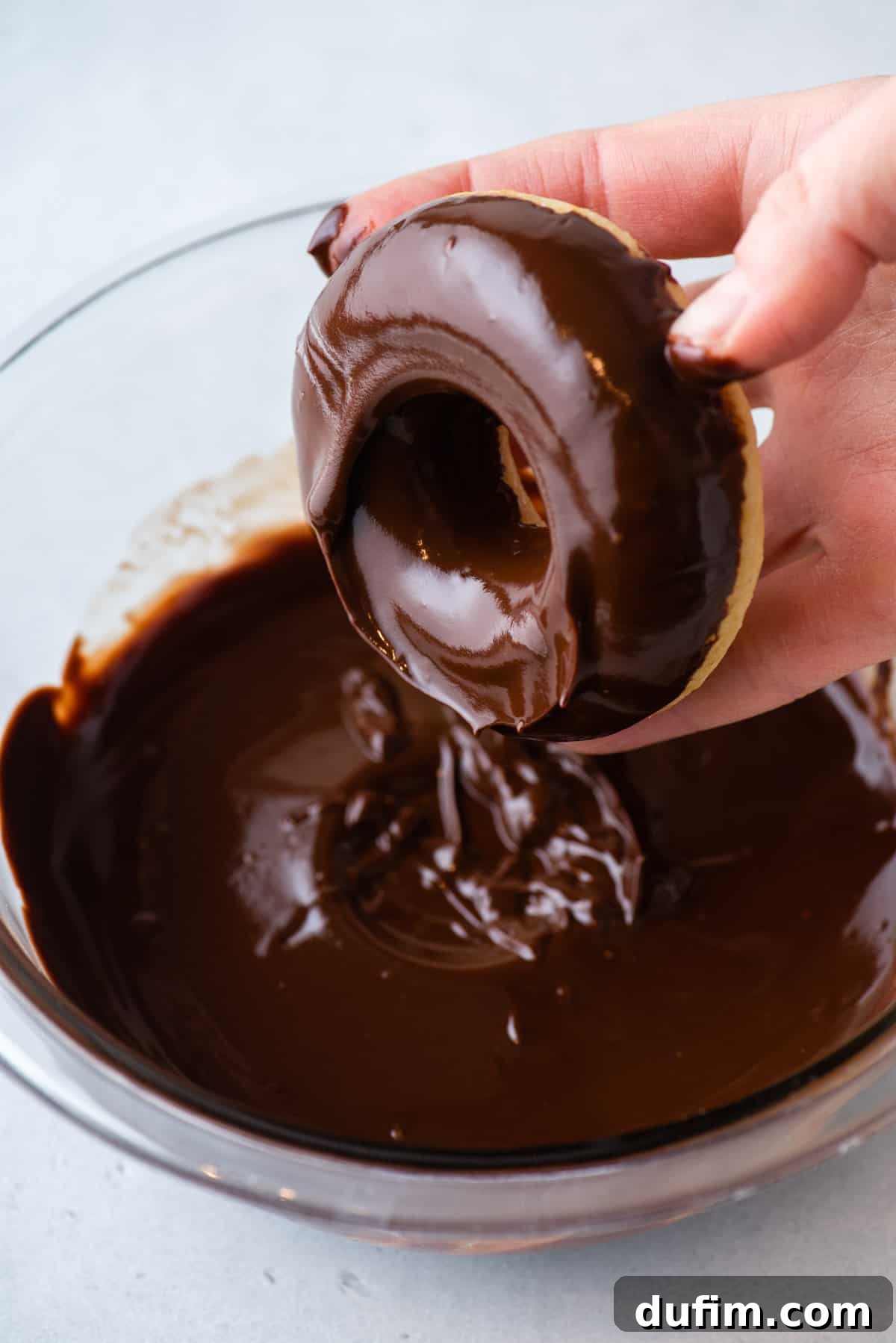 A freshly dipped donut is held above a clear glass bowl filled with chocolate glaze, showcasing the smooth, excess glaze dripping off before setting.