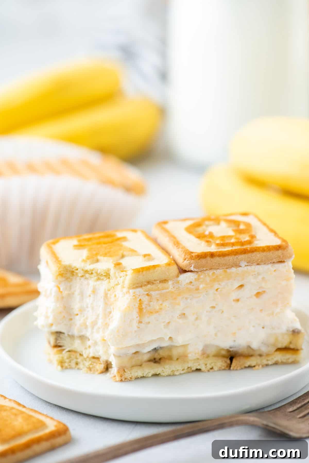 A generous serving of Paula Deen banana pudding on a white plate with a fork, garnished with fresh banana slices and extra Chessmen cookies around it.