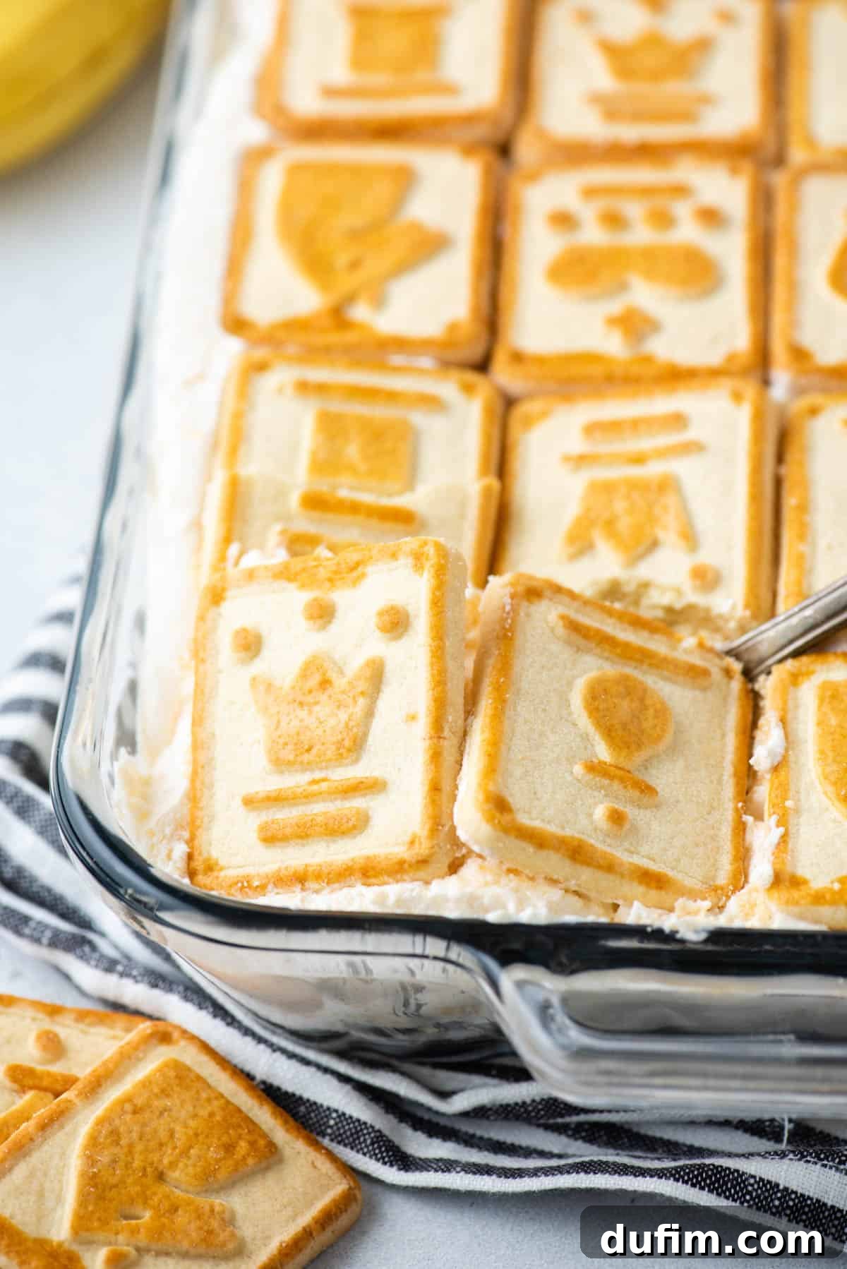 Close-up shot of Paula Deen banana pudding in a clear glass dish, resting on a black and white striped kitchen towel, with Pepperidge Farm Chessmen cookies artfully arranged nearby.