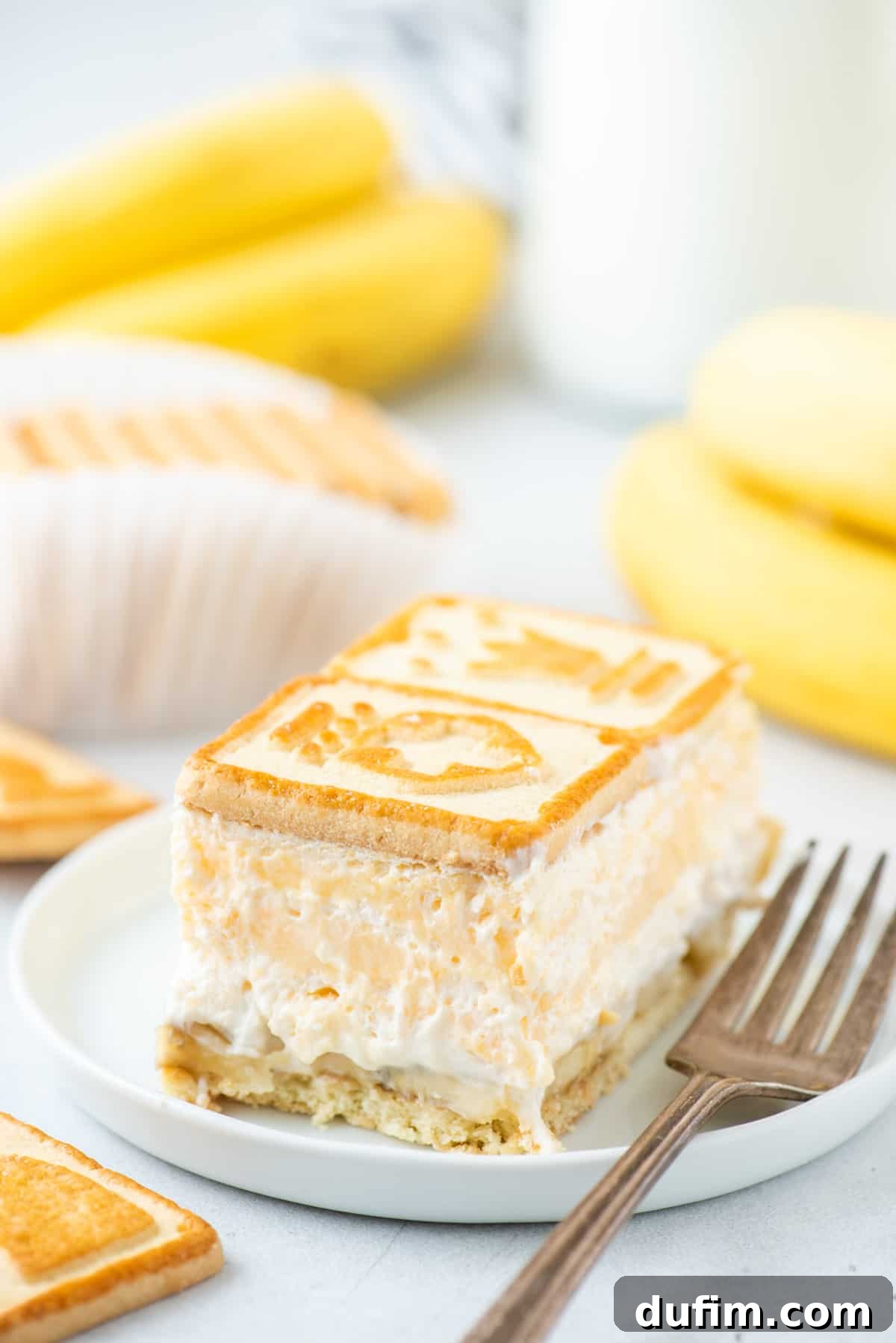 A beautifully presented serving of Paula Deen banana pudding on a pristine white plate, accompanied by a fork and adorned with fresh banana slices and extra Chessmen cookies for visual appeal.