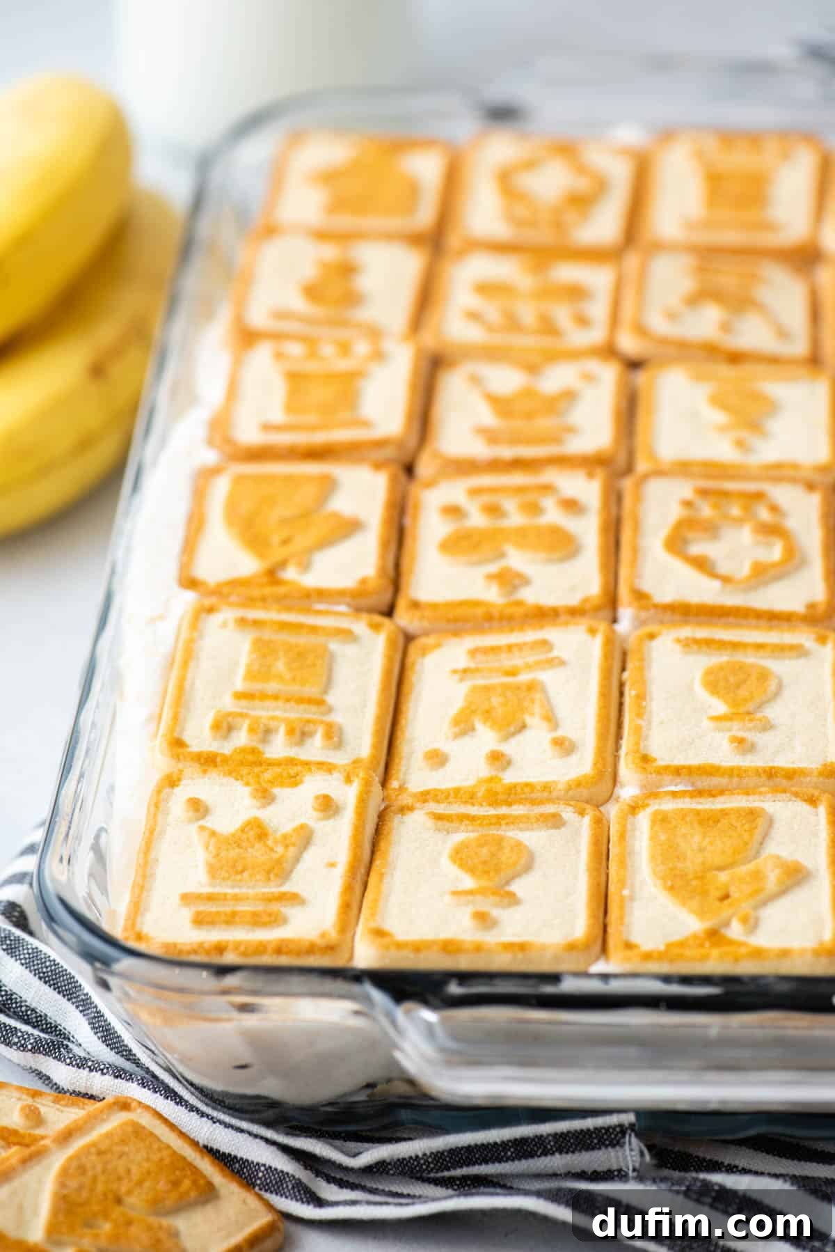 A close-up view of Paula Deen banana pudding showcased in a clear glass dish, resting elegantly on a black and white striped kitchen towel, with additional Chessmen cookies artistically placed around it.