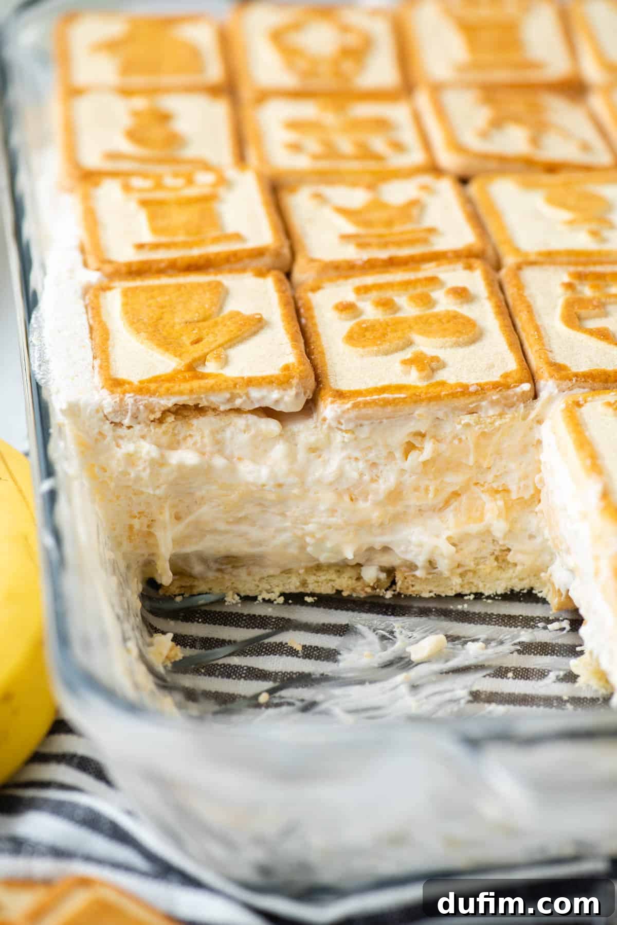 Close-up of a partially eaten serving of Paula Deen banana pudding in a clear glass dish, resting on a black and white striped kitchen towel, with a few Chessmen cookies scattered around the dish.