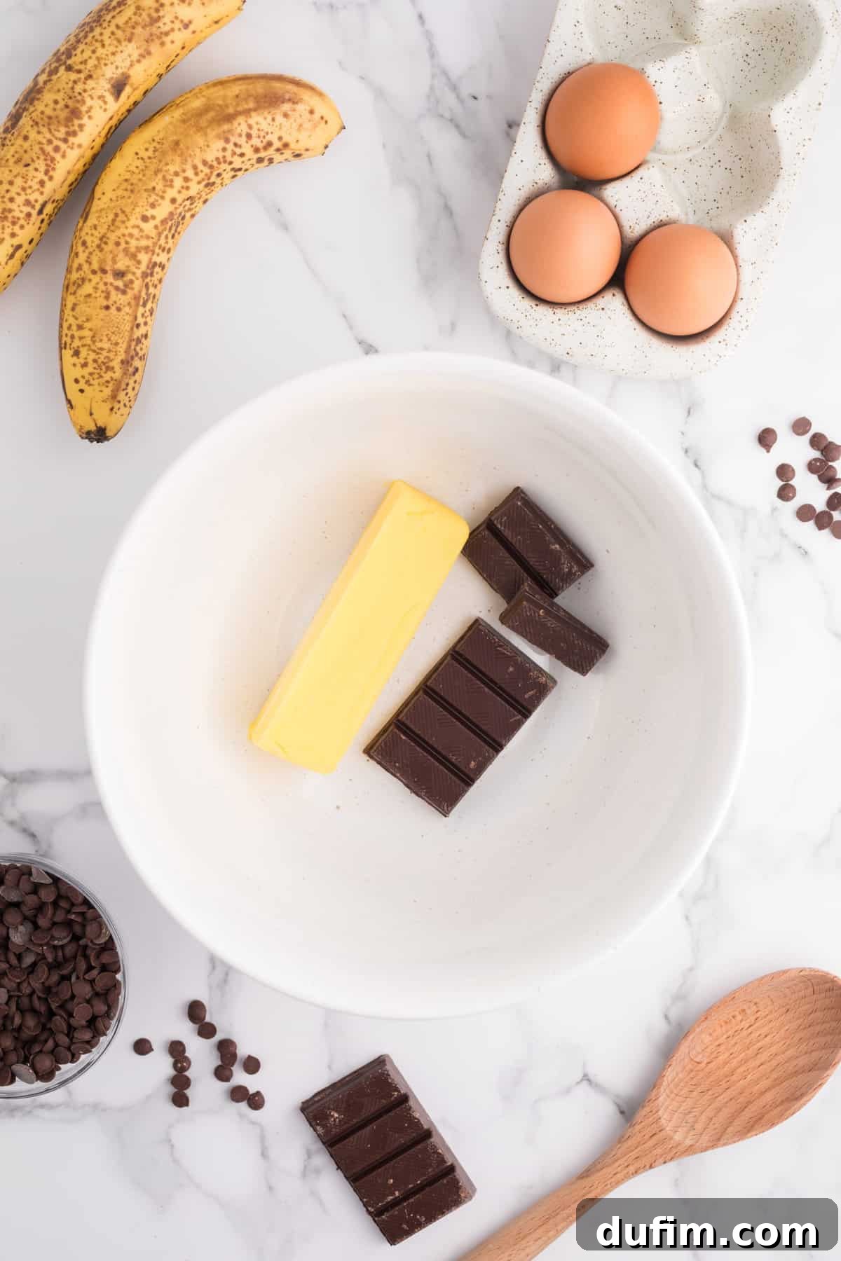 Melted butter and semi-sweet chocolate in a mixing bowl, surrounded by bananas, eggs, a wooden spoon, and chocolate chips – a visual guide to the initial steps of the brownie preparation.