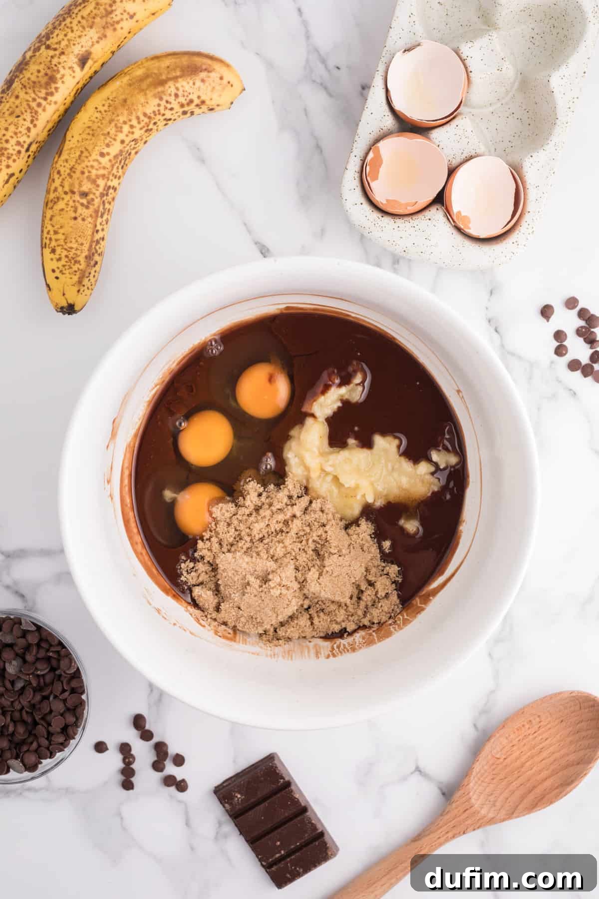 A rich mixture of butter, chocolate, eggs, and brown sugar in a bowl, with ripe bananas, eggshells, a wooden spoon, and additional chocolate ingredients scattered around – illustrating the wet ingredients being combined.