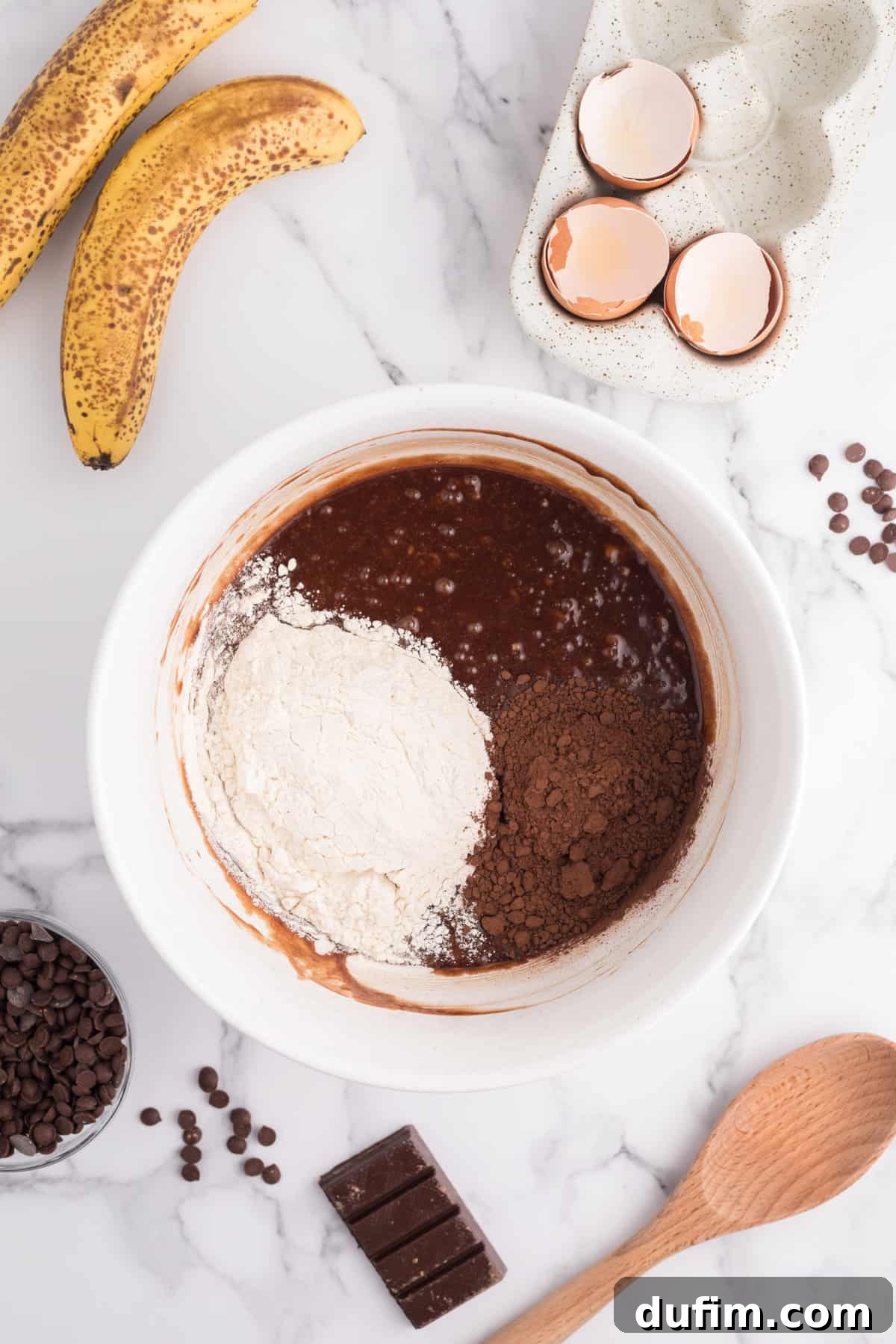 A close-up of the partially mixed banana brownie batter in a white bowl, showing its thick texture and hints of chocolate, with bananas, eggshells, and chocolate chips in the background.