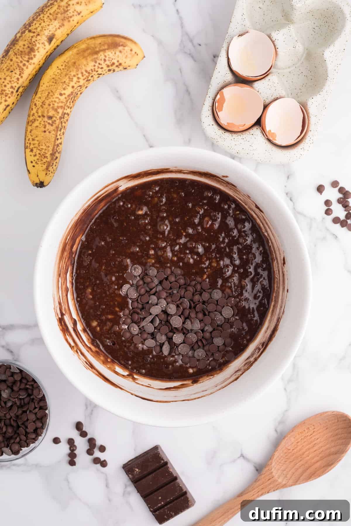 The fully combined banana brownie batter, smooth and ready for baking, in a white bowl, framed by ripe bananas, eggshells, and a scattering of chocolate chips, highlighting the readiness for the oven.