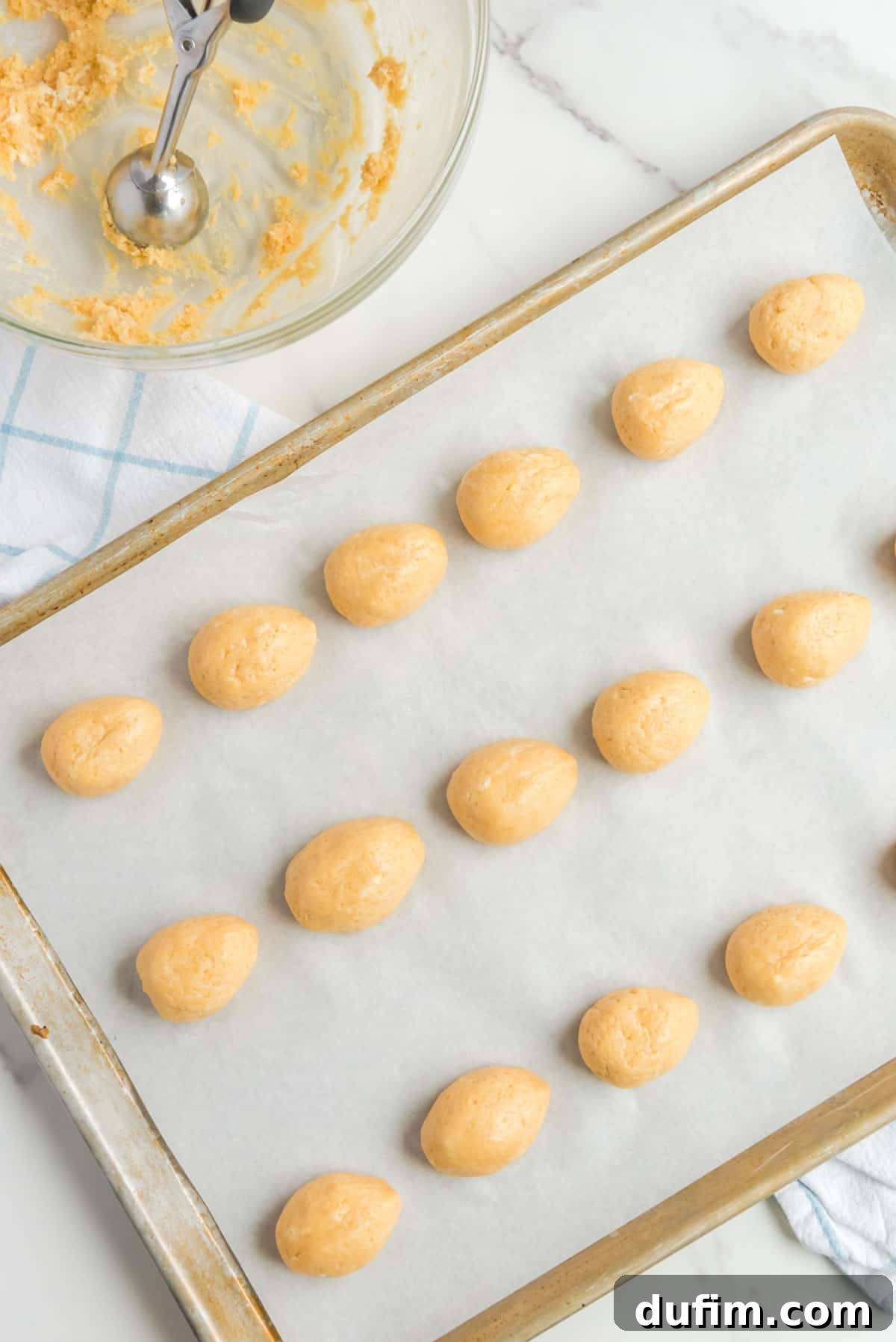 oreo cookie balls shaped into egg shapes, lined up on a baking sheet lined with parchment paper with an almost empty bowl of oreo mix and a cookie scooper beside it