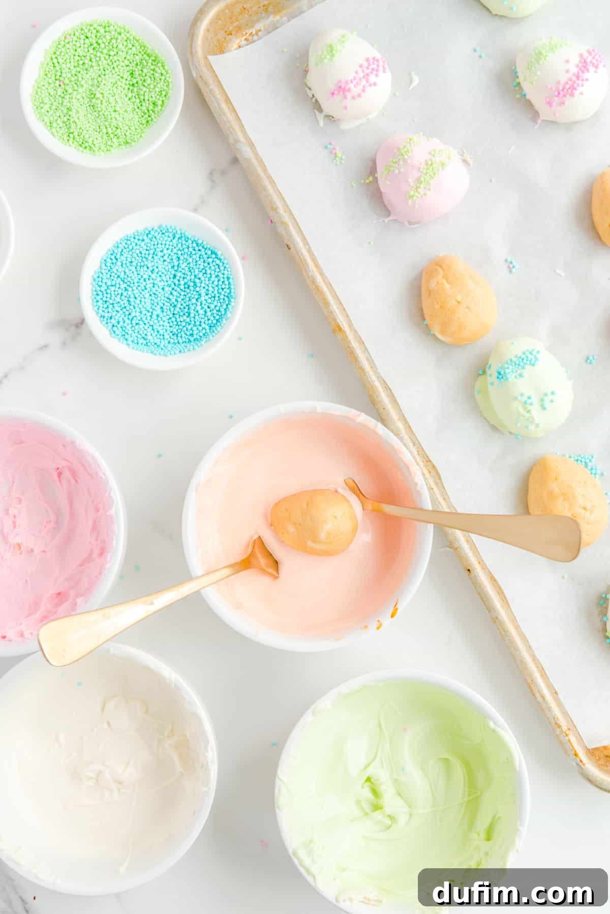 a baking sheet with some dipped and some non-dipped easter egg truffles beside bowls of blue and green sprinkles, and bowls of pink, light orange, light green and white chocolate, with one truffle being dipped with forks into the light orange chocolate.