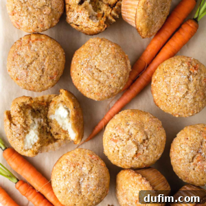 Carrot Cake Muffins are arranged on parchment paper, with a few fresh whole carrots scattered around. One muffin is split open, revealing a creamy filling inside and a speckled, golden-brown top.