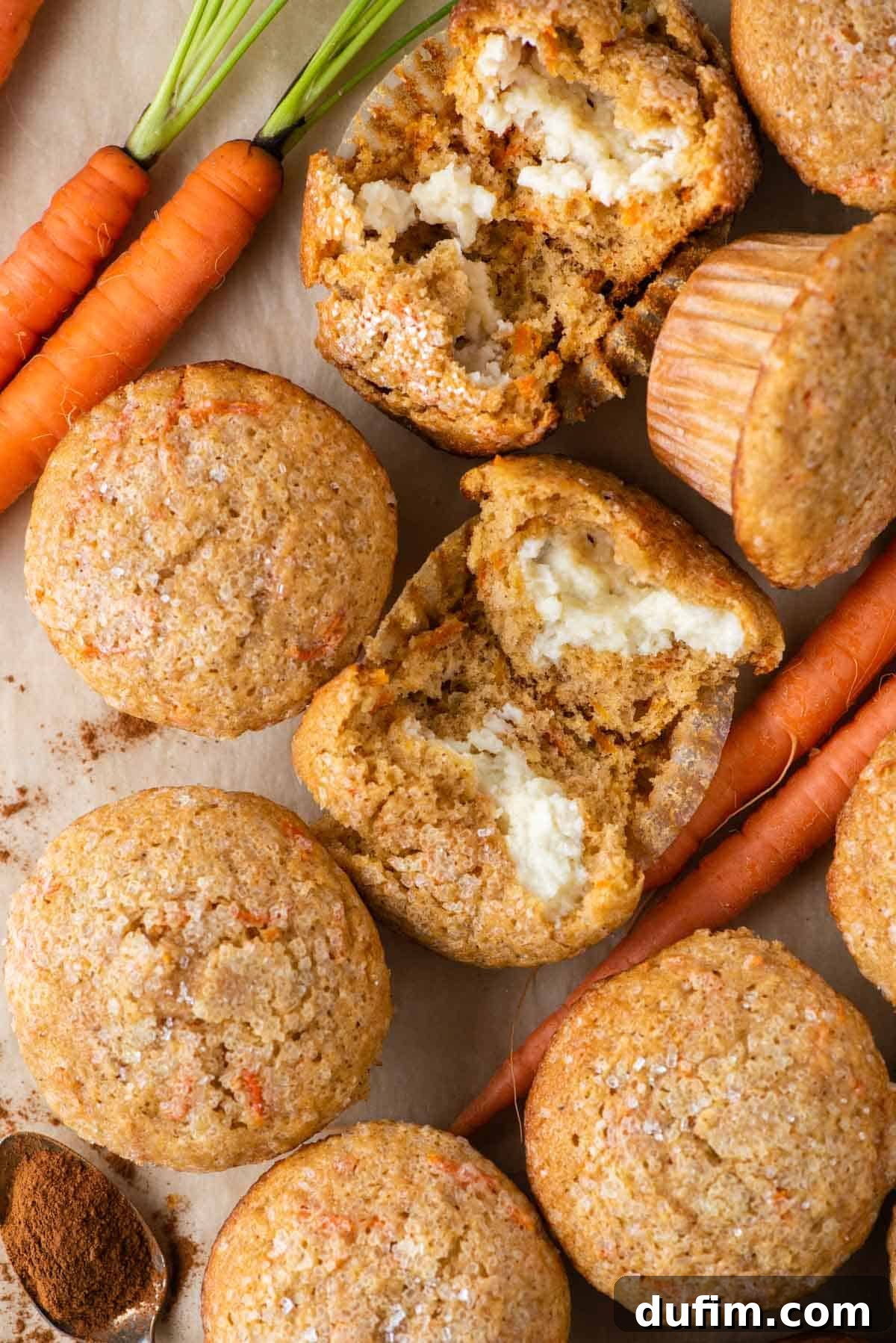 Irresistible Carrot Muffins 2 Carrot cake muffins arranged around carrots, a spoon of spices, and one muffin broken open to reveal the cream cheese filling inside