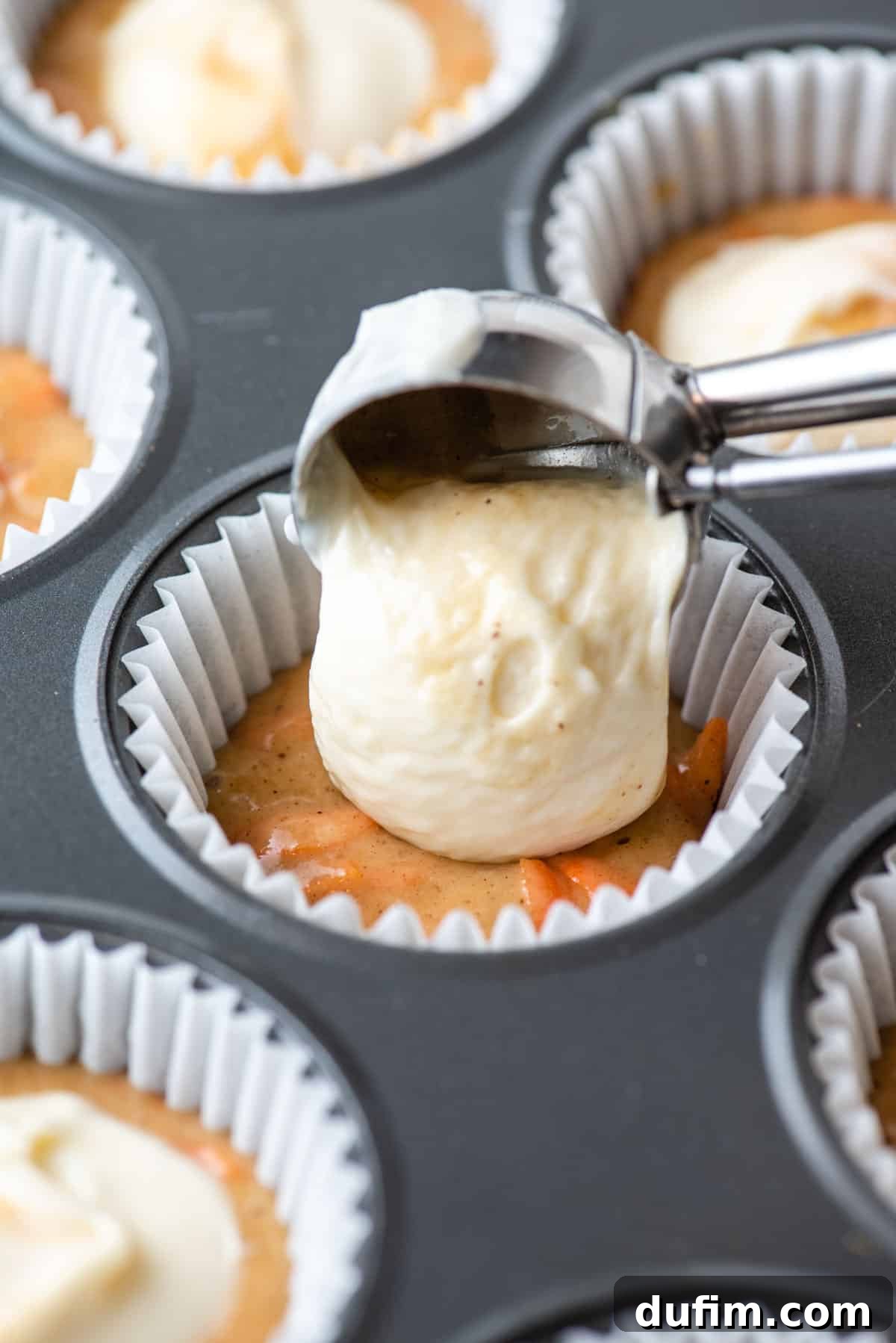 Irresistible Carrot Muffins 7 Carrot cake muffin batter in the bottom of a paper liner in a muffin pan with cream cheese filling being placed on top of it