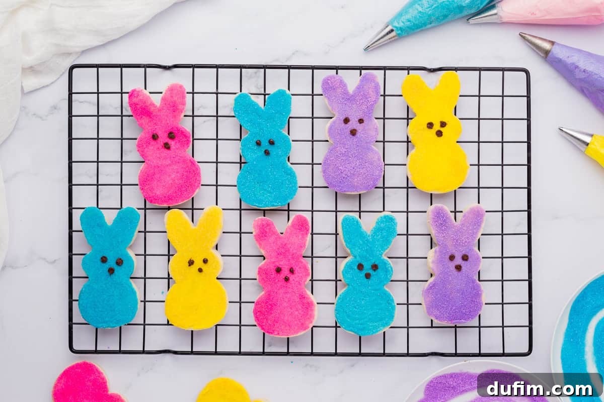 Festive Peeps Bunny Sugar Cookies 2 peeps bunny cookies lined up in two rows on a cooling rack, surrounded by piping bags with blue, pink, purple and yellow icing and bowls of purple and blue sugar