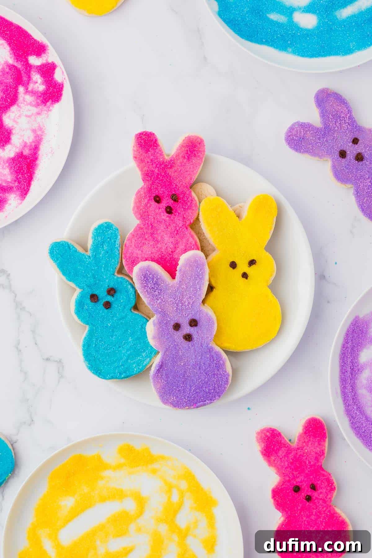 Festive Peeps Bunny Sugar Cookies 3 pink, yellow, blue and purple peeps bunny sugar cookies on a white plate, surrounded by bowls of colored sugar and more cookies