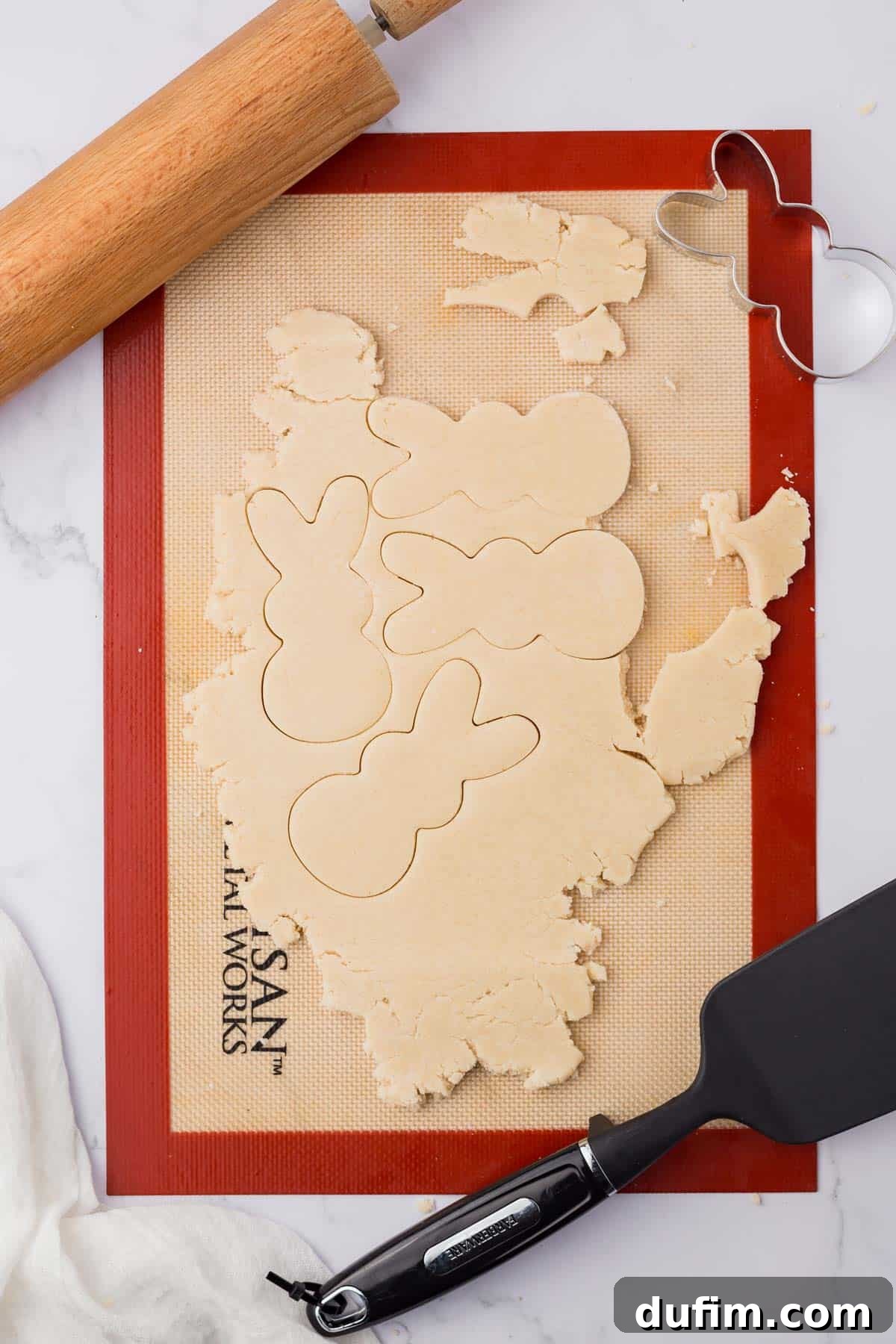 Festive Peeps Bunny Sugar Cookies 5 silicone mat with sugar cookie dough on top, with bunny cutout prints in it, surrounded by a rolling pin, spatula, and peeps bunny cookie cutter