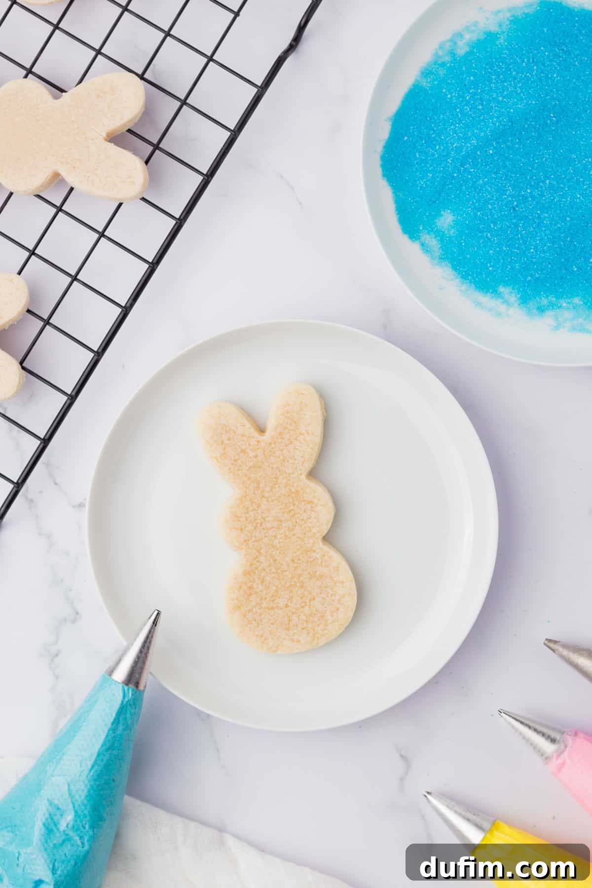 Festive Peeps Bunny Sugar Cookies 6 an undecorated bunny sugar cookie on a white plate surrounded by a bowl of blue sugar, piping bags with colored icing and cooling rack with more cookies