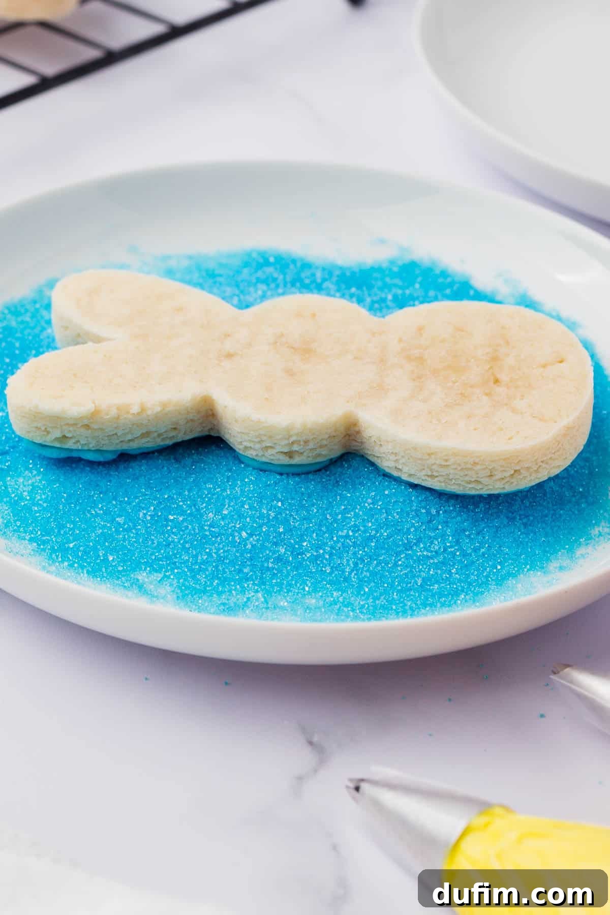 Festive Peeps Bunny Sugar Cookies 7 a peeps bunny sugar cookie with blue frosting on it being dipped in blue sugar on a white plate